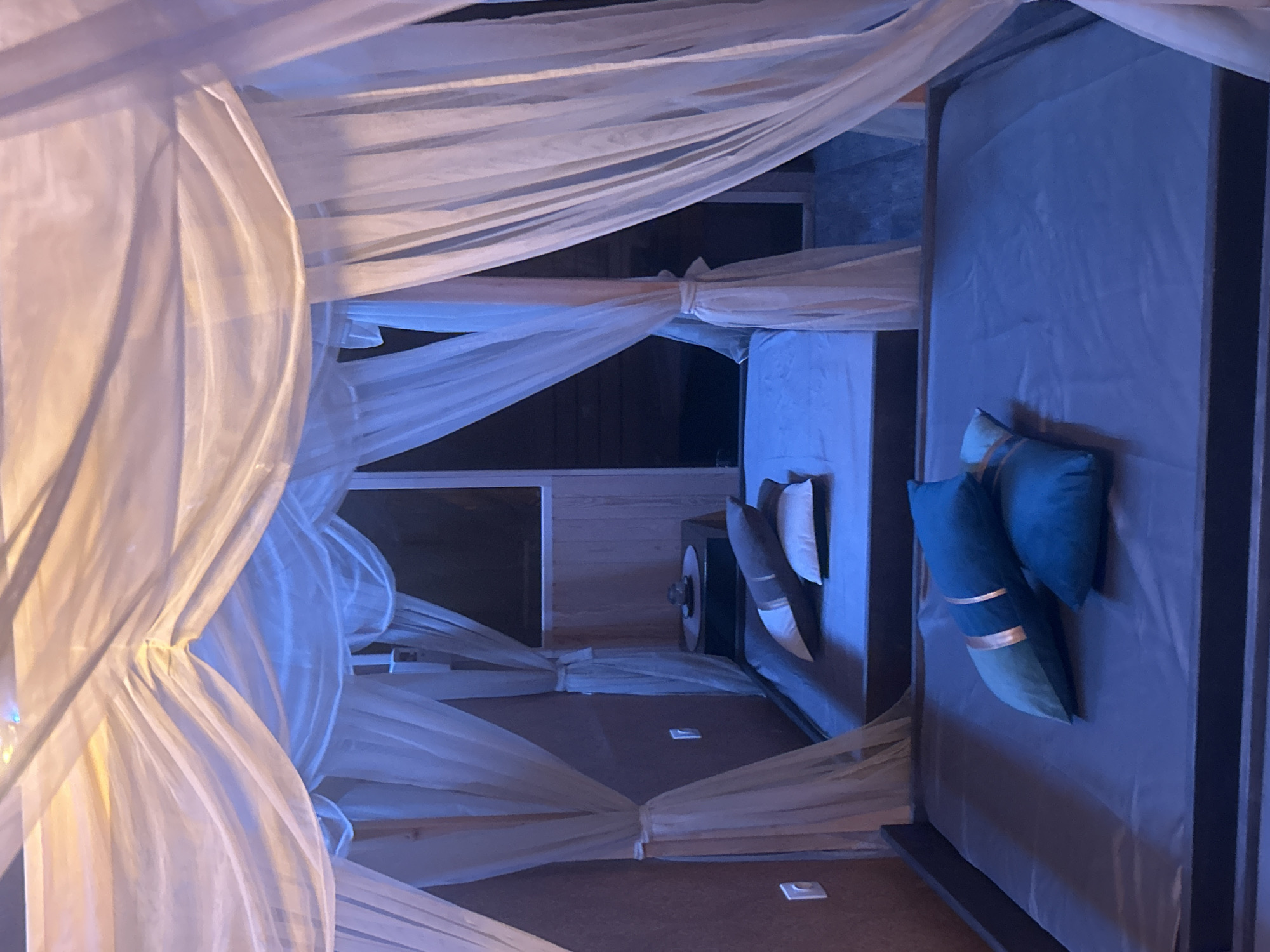 A bedroom features a bed draped with white, translucent fabric, creating a canopy-like effect. The bed is positioned near a window and against a wall. Pillows of various sizes and colors are arranged on the bed. The room appears to be dimly lit. The location is unknown and cannot be determined from the image.
