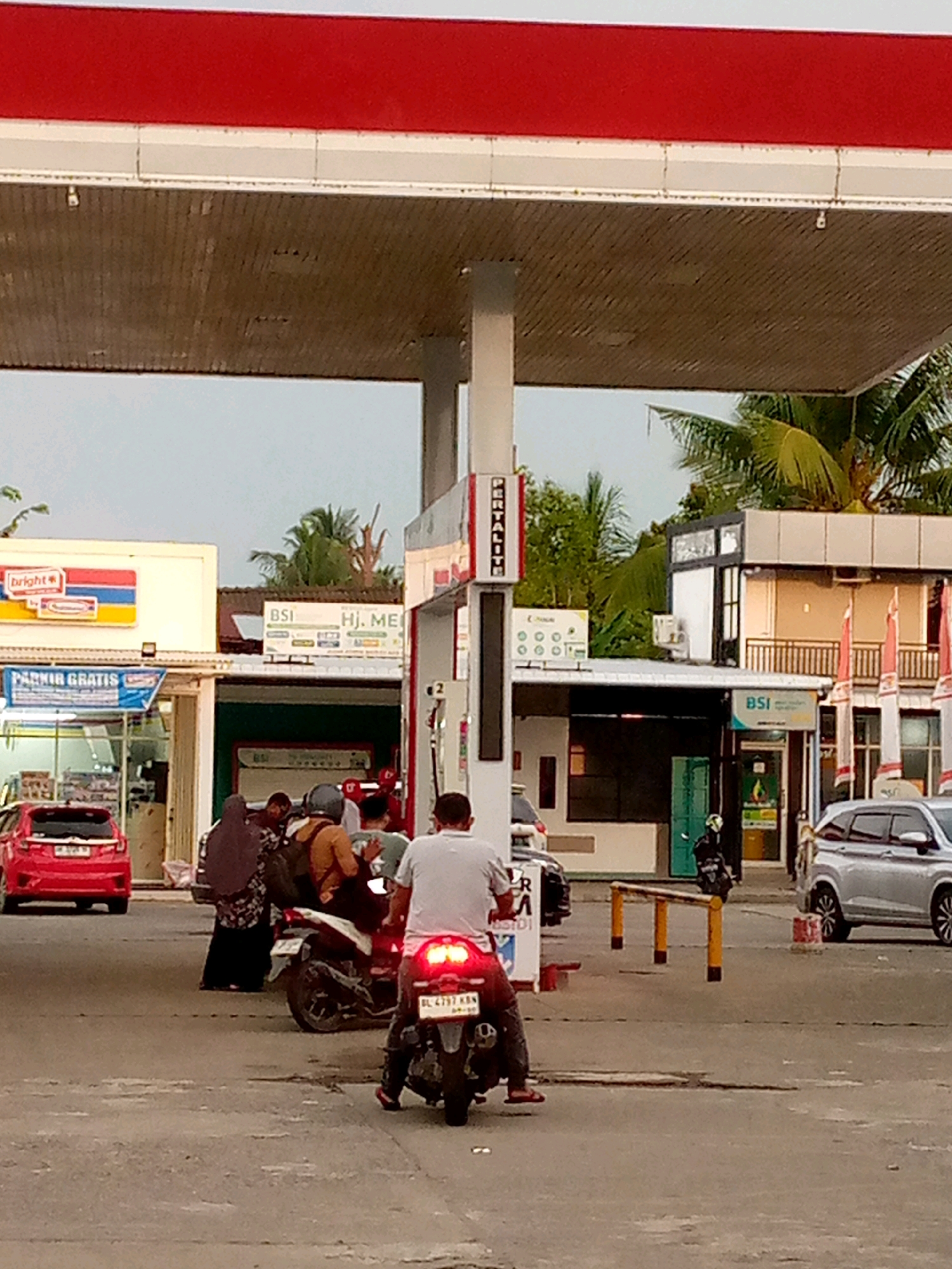 The image depicts a street scene at a gas station in Keudeteupinpunti, Indonesia. A red car is parked to the left, and a silver SUV is on the right. Several people are on motorcycles in the middle of the scene. One motorcycle is occupied by two individuals, one of whom appears to be a woman wearing a hijab. Another motorcycle is ridden by a man in a white shirt and dark pants. A vertical sign with "PERTALITH" in white text on a red background stands near the center of the image. Buildings with awnings and signage are visible in the background. The sky is overcast.