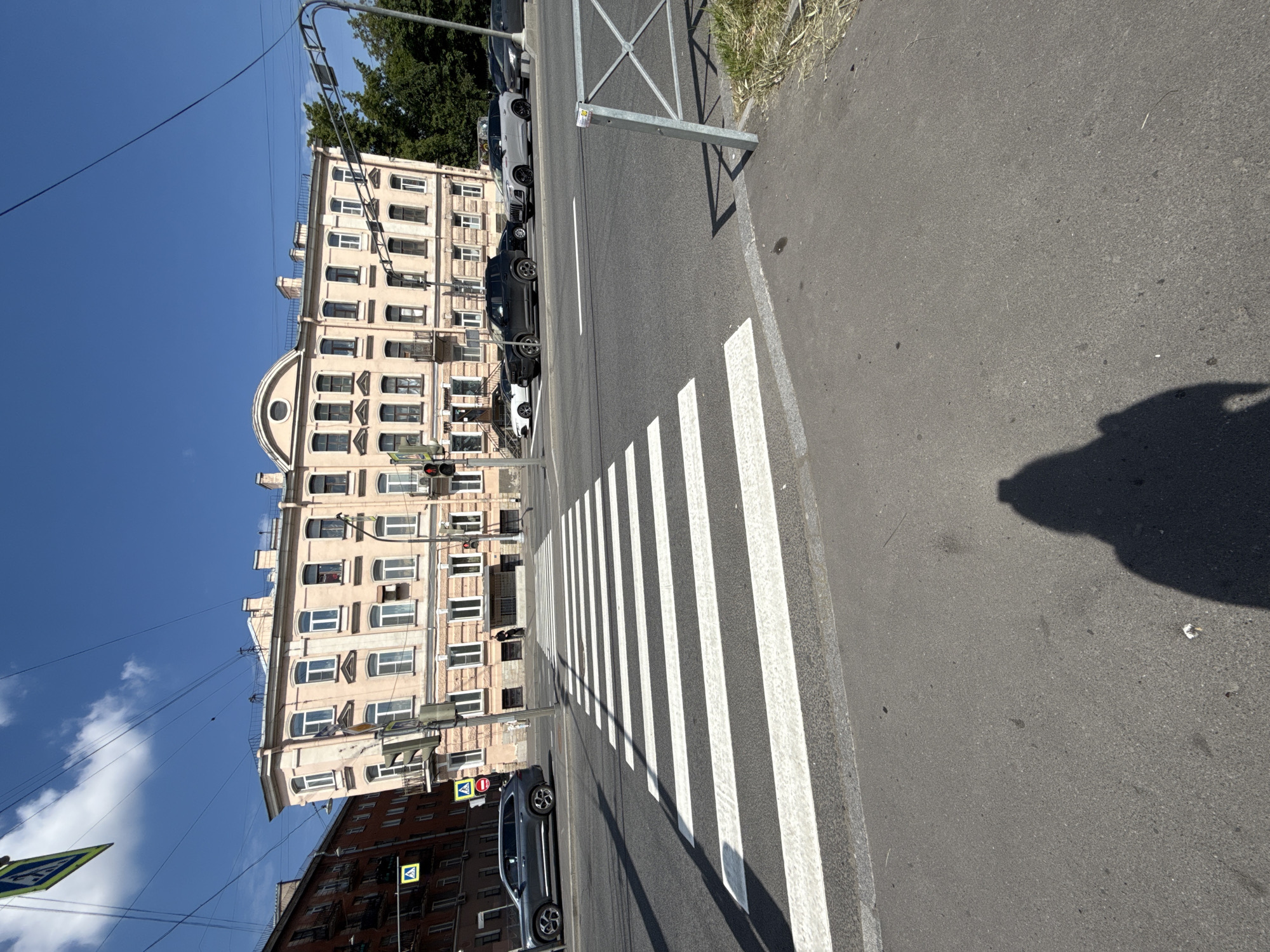 The image depicts a sunny urban street scene, viewed from a slightly elevated perspective looking across a road. The scene is dominated by a wide zebra pedestrian crossing (crosswalk) stretching across an asphalt road in the foreground and midground.
On the left side of the road, a large, multi-story building with a light, possibly beige, facade stands prominently. This building features many windows and an distinctive arched architectural element at its lower level. Several cars, including a white and a dark SUV, are parked along the curb in front of this building. Another dark gray SUV is visible on the street, near the pedestrian crossing. Behind the buildings, green trees are visible, suggesting nearby foliage.
To the right of the road, there is a paved sidewalk bordered by a metal barrier fence. Dry grass can be seen along the edge of the sidewalk. Long shadows are cast across the road and sidewalk by the fence and an unseen object or person, indicating bright sunshine and possibly morning or late afternoon.
The sky is clear and blue with a few wispy white clouds. Overhead wires, traffic lights (one showing a red light for the street ahead), and street light poles are visible. Several road signs are present, including blue square signs with a white triangle and black pedestrian symbol, indicating a pedestrian crossing. A small, unreadable yellow sticker is attached to the metal barrier fence. No people are explicitly visible, and no specific activity is taking place beyond the general urban setting.