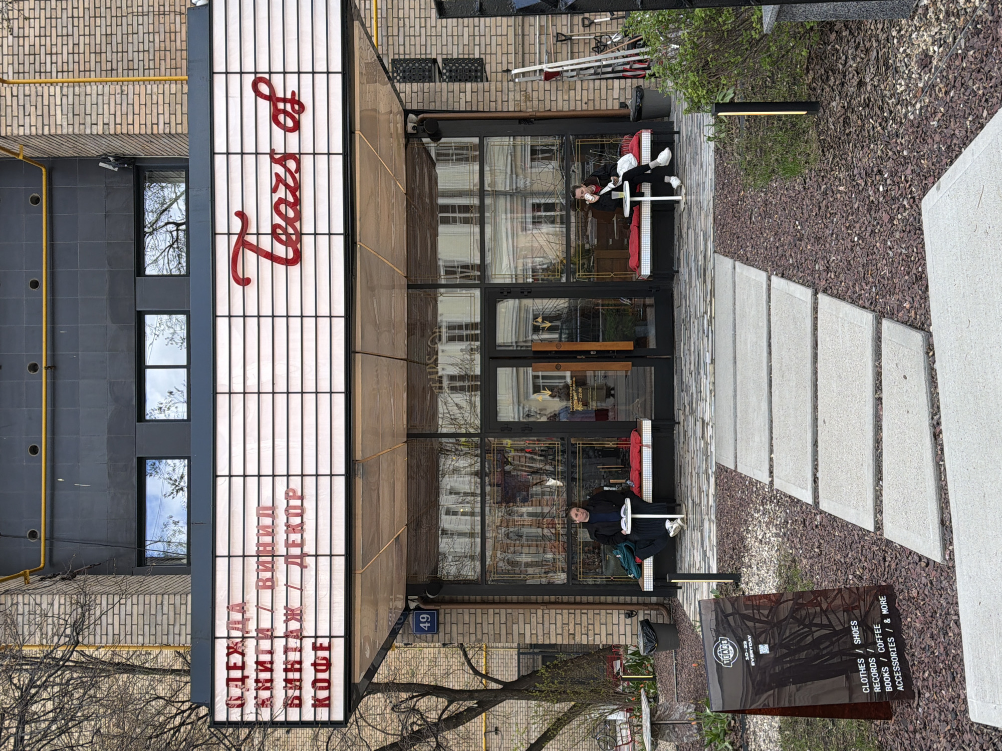 This image captures the facade of a shop or cafe in Moscow, Russia, featuring an entrance with outdoor seating under an overcast daytime sky.
The building displays a mix of architectural styles, with upper levels constructed from light brown brick and the lower section covered in dark gray tiles. A prominent, vertically oriented illuminated sign above the entrance reads "Tears of" in red cursive script. Below this, in red block letters, are the Russian words "ОДЕЖДА / ОБУВЬ" (Clothes / Shoes), "ВИНИЛ / ДЕКОР" (Vinyl / Decor), and "КНИГИ / КОФЕ" (Books / Coffee). The building number "49" is affixed to the facade near the entrance.
Two individuals are seated at small, round white tables with red chairs, positioned outside the glass-fronted entrance. One person, dressed in dark attire, is looking towards the viewer, while the other is looking away. The glass reflects the street and adjacent structures.
The ground in front of the building is covered with a bed of dark reddish-brown mulch, interspersed with some green foliage. A path of rectangular light gray concrete stepping stones leads away from the entrance. An A-frame sign near the path provides additional information, stating "TEARS 10-22 EVERYDAY" and listing categories of items: "CLOTHES / SHOES", "RECORDS /", "BOOKS / COFFEE", and "ACCESSORIES / & MORE". A yellow gas pipe runs vertically along the left side of the brick building. The scene depicts a casual, urban commercial setting.