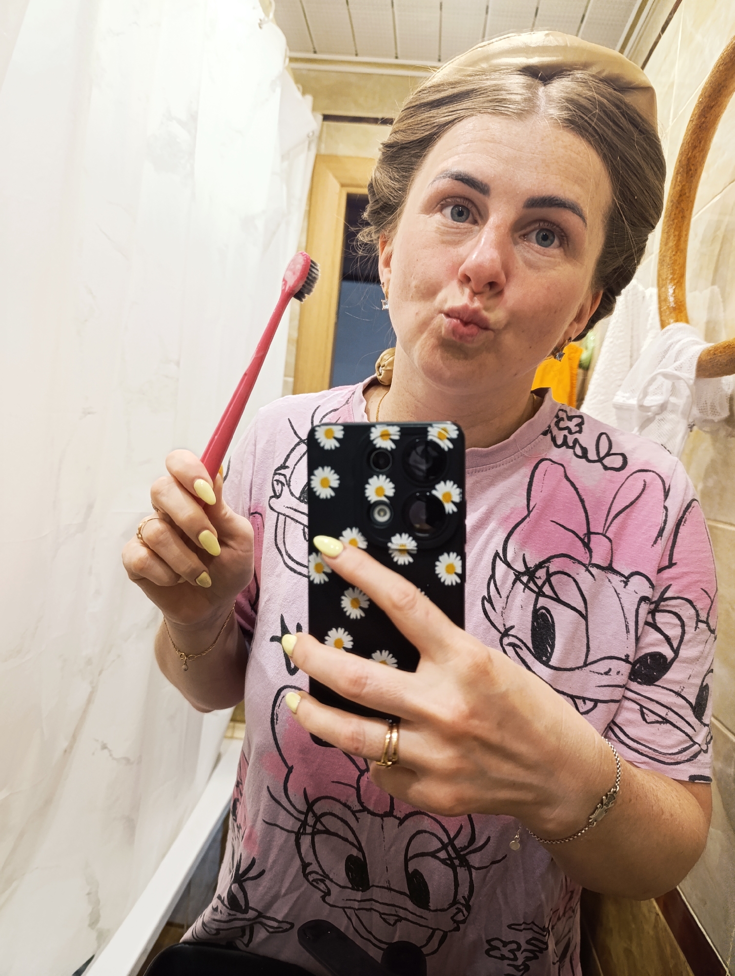 The image shows a person in a bathroom, possibly taking a selfie. The person is holding a toothbrush in one hand and a phone in the other, which has a daisy-patterned case. The person is wearing a pink t-shirt with a cartoon character on it and has yellow nail polish. They appear to be making a kissing face. The background shows a bathroom setting, including a shower curtain, tiled walls, and a mirror. The specific location in the city of Unknown, Unknown is not discernible from the image.
