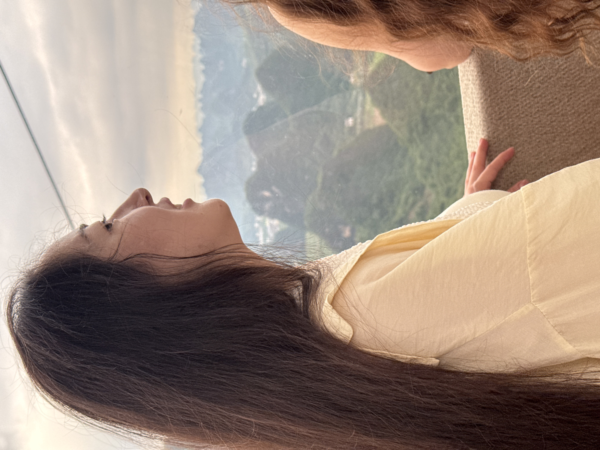 A woman with long, dark hair, wearing a light yellow shirt, leans against a cushioned surface. A second person with brown hair is partially visible in the upper right corner. The background reveals a hazy vista of hills and vegetation. Based on the landscape, the location is likely Guilin, China, where similar scenery is common. The woman is looking up, seemingly observing the view. The presence of glass or some transparent barrier suggests the scene is inside a structure with an observation point.