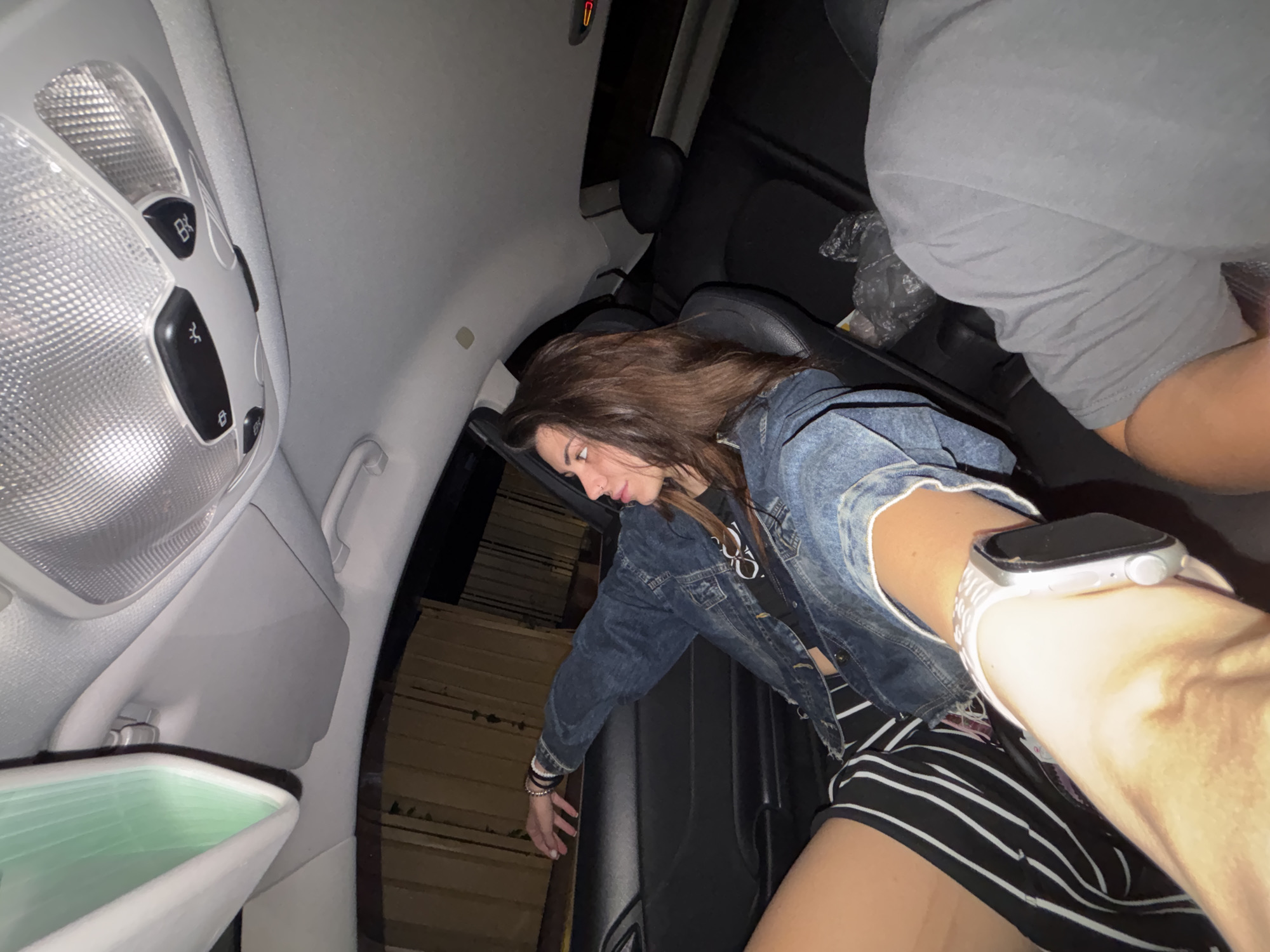The scene depicts the interior of a Mercedes-Benz vehicle, possibly a C-Class or CLK model, at night, located in the city of Sofia, Bulgaria. Illumination within the vehicle is provided by its overhead dome lights.
In the left-center, a young woman is seated in a black leather-upholstered car seat. She has long, dark brown hair and a visible nose piercing on her right nostril. She wears a light blue denim jacket over a dark top, paired with black and white striped shorts or a skirt. Her left arm is casually extended, resting on the black door panel. Her right forearm, equipped with a silver-colored Apple Watch featuring a white band, is prominent in the immediate foreground, suggesting she is closer to the camera. She is looking towards her left, away from the camera's direct line of sight.
In the bottom-right foreground, a portion of another individual is visible, comprising a shoulder and upper arm, clad in a grey t-shirt. This person is holding the camera.
The car's interior features a light grey fabric headliner and an overhead console housing two textured, clear plastic dome lights and black control buttons, including light activation and door-controlled light functions. A partial view of a rearview mirror with a greenish reflection is visible in the upper-left. Through the window on the left, an exterior view reveals a wooden slatted fence or wall, dimly lit from above, indicating a stationary position in an urban or suburban environment. The rear passenger seats are empty, with a black plastic bag visible on one.