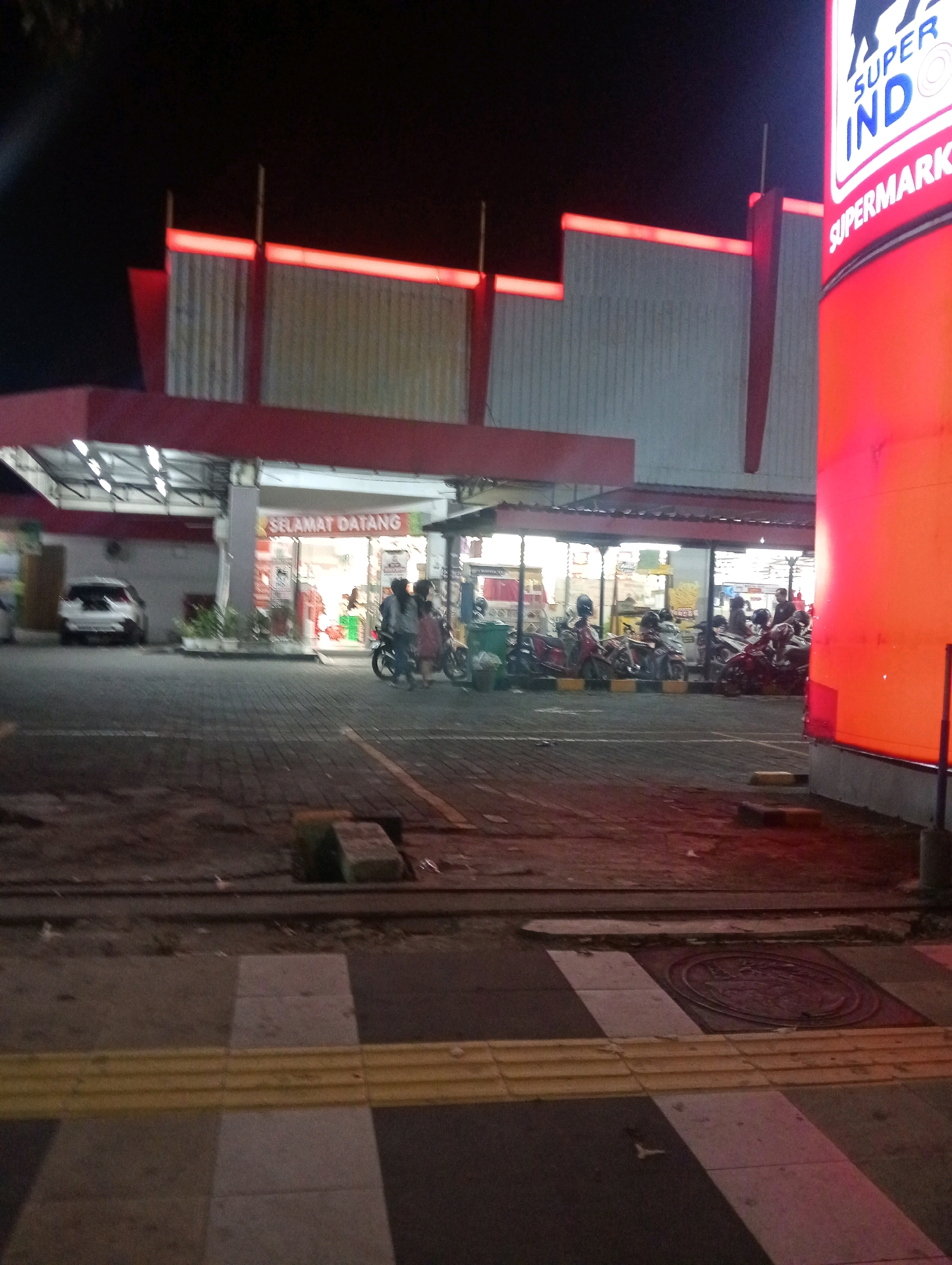 The image captures the exterior of a Super Indo supermarket in Bojonegoro, Indonesia, at night. A large, illuminated red sign with white text advertising "SUPER INDO SUPERMARKET" is prominent on the right side of the frame. The supermarket building features a red awning over the entrance area, which is lit, revealing shelves stocked with products inside. A white car is parked to the left of the entrance. In front of the supermarket, a row of motorcycles is parked in a paved area. Several individuals are present near the entrance, with some standing and others near motorcycles. The ground in front of the supermarket is paved with dark bricks, and a pedestrian crossing with a yellow tactile paving strip is visible in the foreground. The overall environment suggests a commercial area active in the evening.