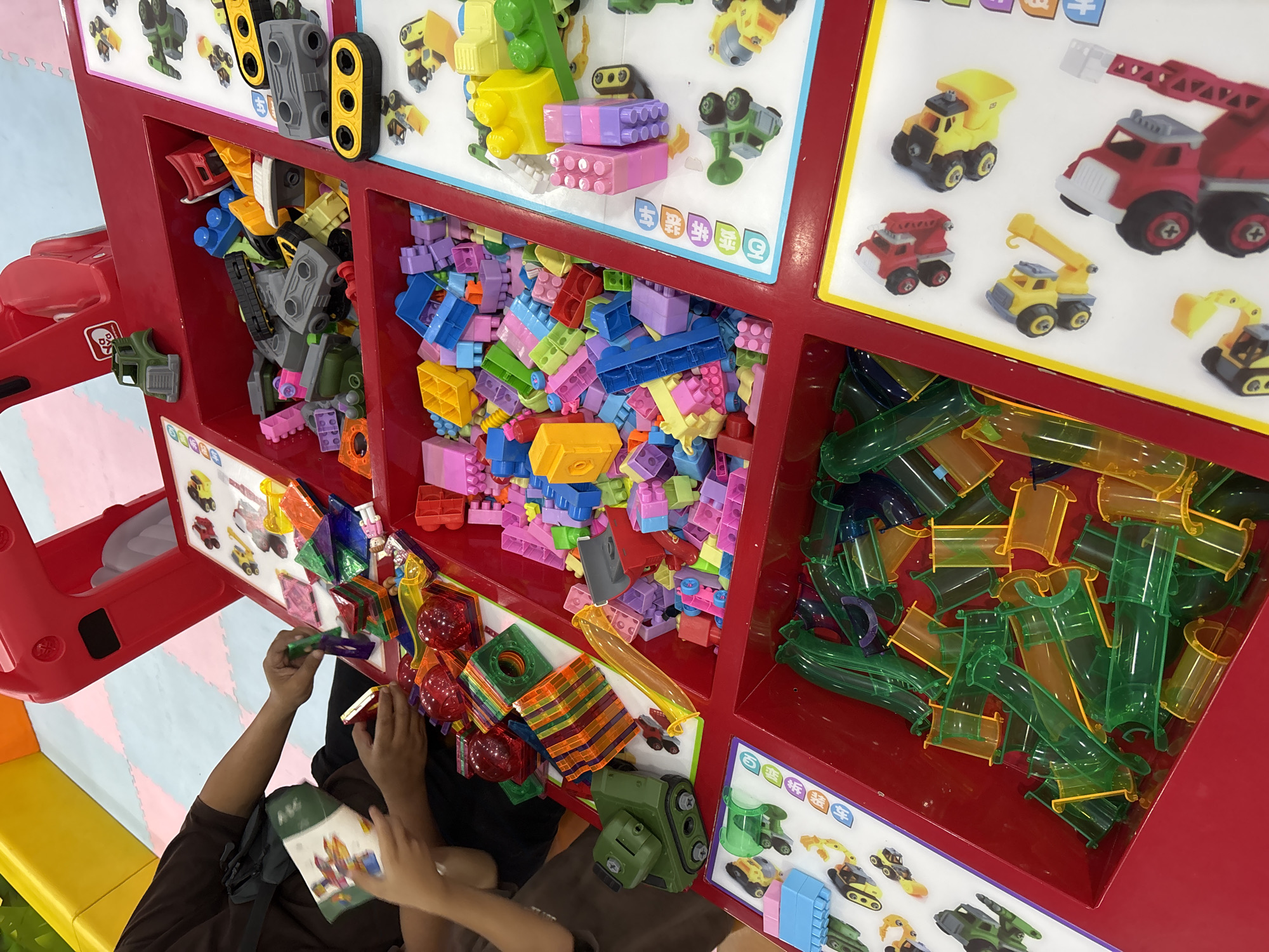 In a brightly lit indoor play area in Liangjulang, Indonesia, a close-up shot captures a collection of colorful children's toys. The toys are predominantly made of plastic and are sorted into red compartments of a large, multi-tiered play structure. One person's hands are visible, engaged with a book and some magnetic building blocks in the top left section of the image. The toys include a variety of building blocks in assorted colors and shapes, transparent tubes for marble runs or construction, toy vehicles such as tanks and construction equipment, and magnetic geometric shapes. The background shows the soft, padded flooring typical of a play center, with yellow and pink cushioned barriers visible. The overall scene is one of a well-stocked and engaging play environment for young children.