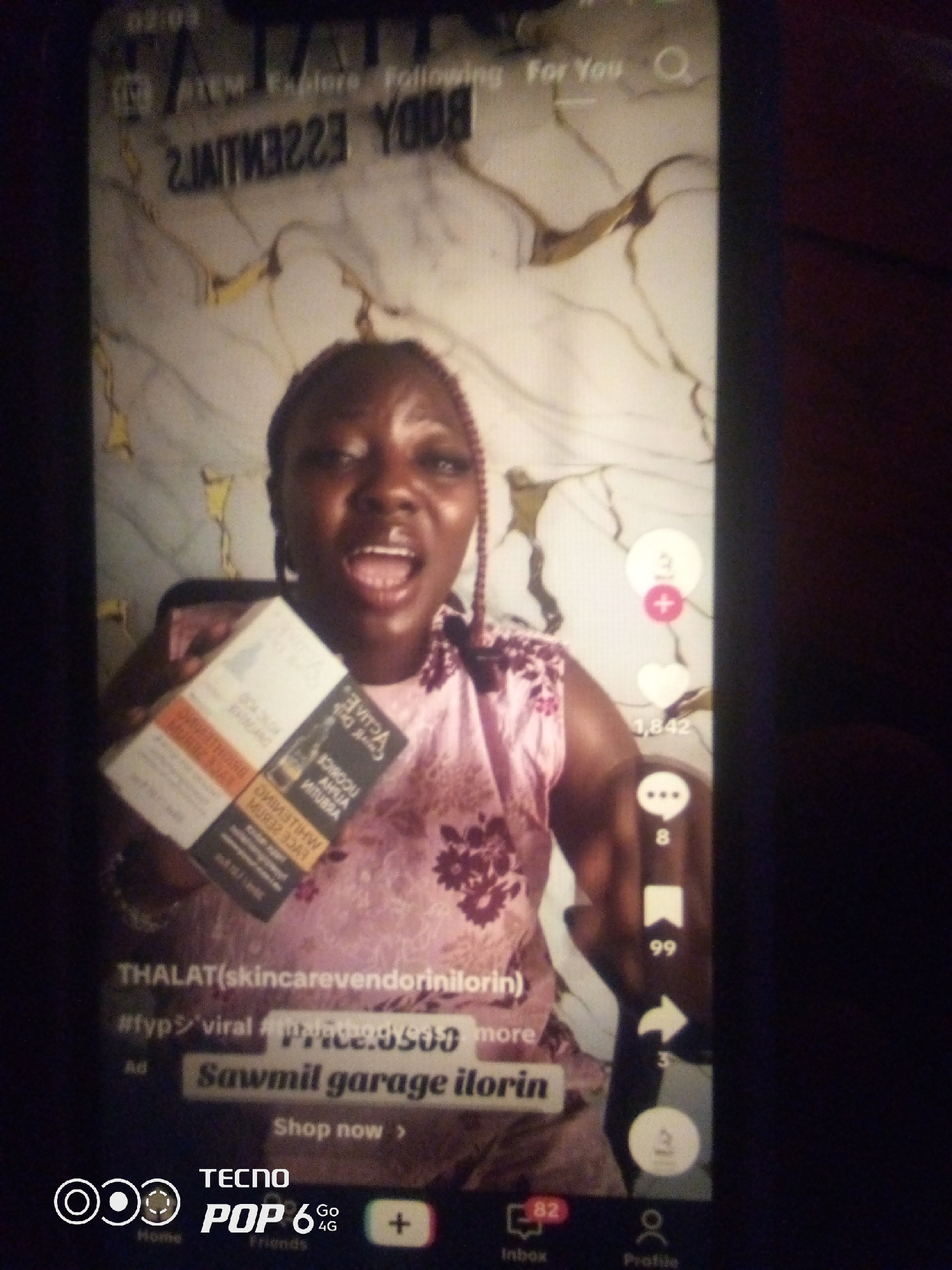 This is a vertical video, likely a social media clip, filmed indoors with a dark, possibly wooden background featuring a marbled or textured pattern. The lighting suggests it's daytime, though no external cues like windows or weather are visible.
The primary subject is a Black woman with braided hair, wearing a pink patterned top. She is holding up two product boxes, angled towards the camera. One box is predominantly white with orange and black text, while the other is black with white and orange text. Her mouth is open, and she appears to be speaking or exclaiming with excitement.
On the right side of the screen, social media interface elements are visible, including interaction icons for likes, comments, and shares, with corresponding numbers. Text overlays in the bottom section of the video provide context: "THALAT(skincarevendorinilorin)", "#fypviral", a price "Price: 3500", and "Sawmil garage ilorin". The Tecno Pop 6 logo and navigation icons for "Home", "Friends", "Inbox", and "Profile" are at the bottom, indicating the platform is likely TikTok. The overall mood is energetic and promotional, likely advertising skincare products.
