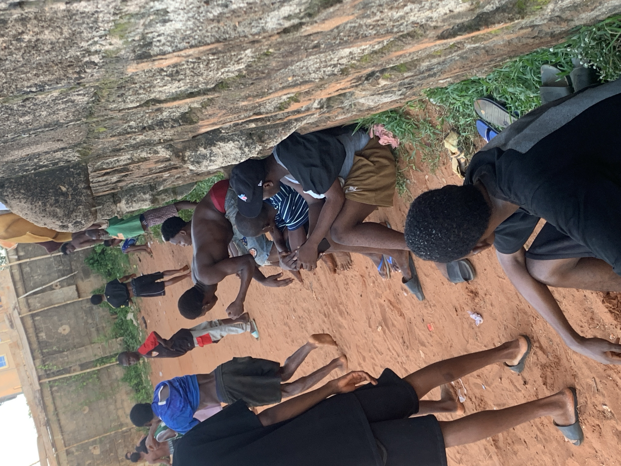A group of approximately fifteen to twenty young males, ranging from boys to young adults, are engaged in an outdoor activity on a reddish-brown dirt ground. The setting is likely Ukwu-Abwa, Nigeria, given the context. Many individuals are in various crouched, kneeling, or prone positions, suggesting a structured physical exercise session, drill, or possibly a communal gathering with elements of physical training or prayer. Several are in push-up or plank-like stances, while others are kneeling with hands clasped or resting on the ground.
Most participants are dressed casually in shorts, t-shirts, or vests, with some individuals shirtless. Footwear varies; some are barefoot, while others wear sandals or slippers. One pair of shorts clearly displays the "NIKE" logo.
The background features a prominent, weathered stone or concrete wall running across the upper portion of the scene. Patches of green grass and sparse vegetation grow around the base of this structure. Further in the distance, partially visible concrete block buildings and more greenery indicate a semi-urban or rural environment. The scene is illuminated by bright daylight, suggesting a daytime activity. The overall impression is one of disciplined group activity or training.
