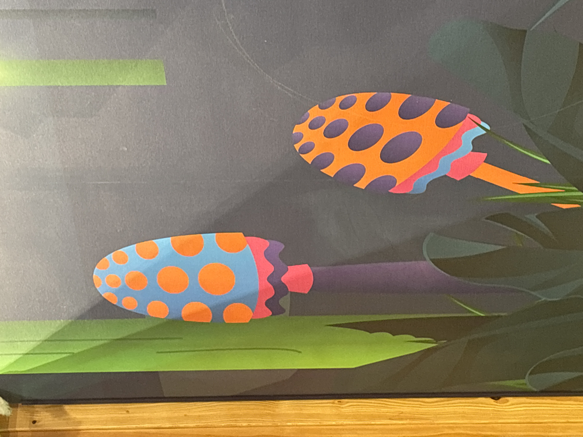 The image shows a close-up of a colorful mural or artwork featuring stylized mushrooms against a dark gray background. To the left, a wooden vertical beam with a visible grain is present. The mural itself depicts two mushrooms with bright orange caps dotted with purple circles. The mushroom on the left has a purple stem and a ruffled, multi-colored base in pink, purple, and light blue. The mushroom on the right is smaller, with an orange stem and similar orange and purple spotted cap, featuring a ruffled base in pink and light blue. Green foliage is depicted at the bottom of the image, surrounding the mushrooms. There are also vertical green stripes painted on the gray background, one of which is partially obscured by a wooden beam on the far left. The overall scene is vibrant and abstract, with no people or discernible actions. The location is not identifiable within the city of Dubai, United Arab Emirates.