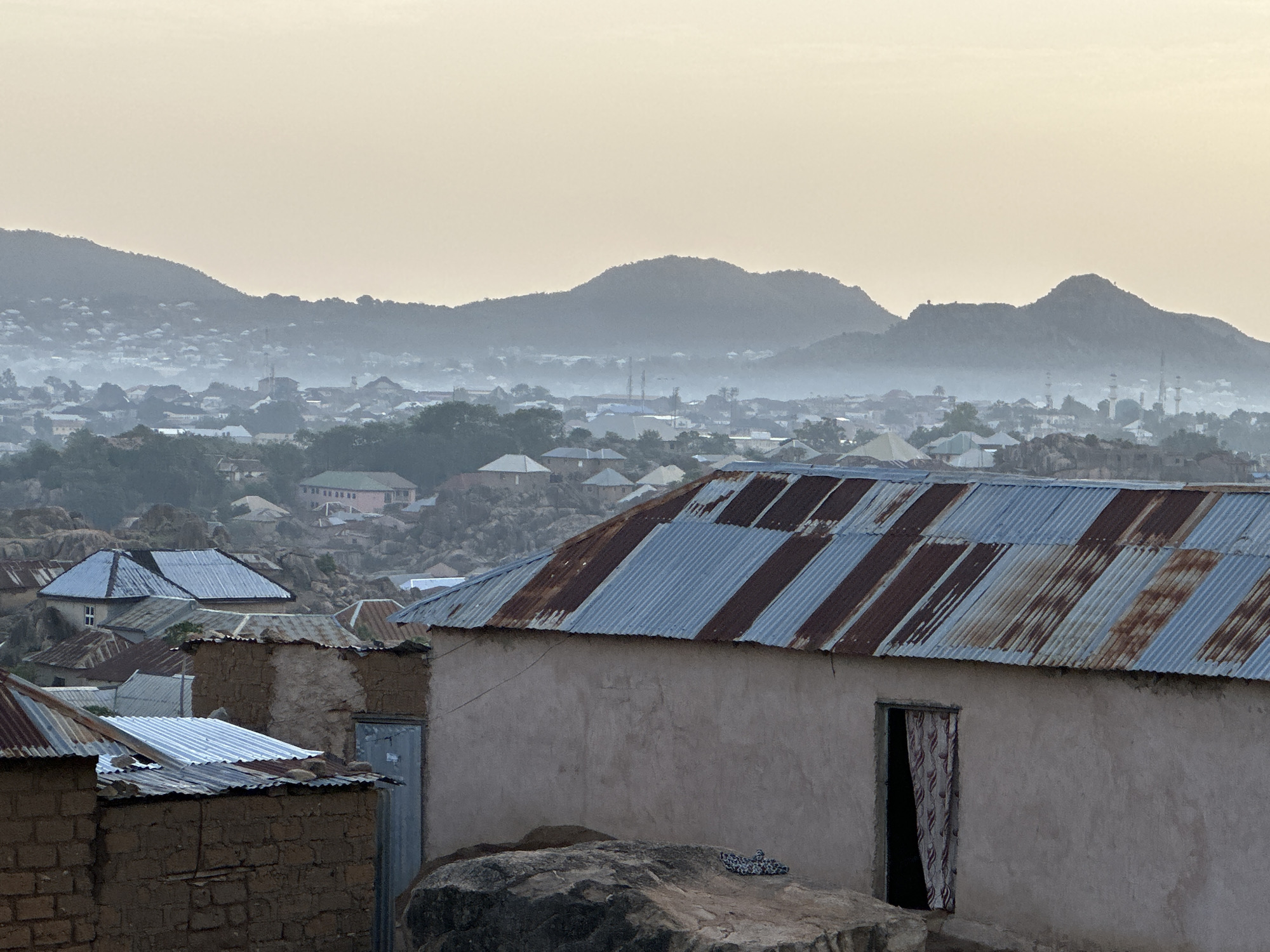 The image depicts a view of Jos, Nigeria, at dawn. In the foreground, a collection of small, modest buildings with corrugated metal roofs sit atop rocky outcrops. The roofs exhibit alternating stripes of silver and rust. The walls of these buildings are constructed from mud or cement and appear weathered. An open doorway reveals a glimpse of a curtain inside. In the midground, the cityscape stretches into the distance, with buildings interspersed among rocky formations and trees. A layer of haze obscures the more distant structures, softening their outlines. Rising behind the city are several mountain peaks, their forms also blurred by the atmospheric conditions. The sky is an even, pale gray, suggesting the early morning light.