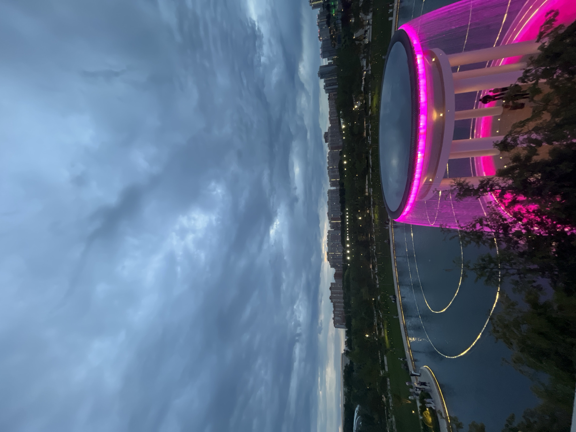 The image displays a captivating night scene in a modern urban park, likely located in Krasnodar, Russia. Above, the sky is dark and heavily overcast with thick grey clouds, indicating either late dusk or an evening with inclement weather. Along the distant horizon, a cityscape unfolds with numerous multi-story buildings, some illuminated, suggesting a vibrant urban setting.
The foreground and mid-ground are dominated by impressive park architecture. On the right, a large, circular water feature, possibly a fountain or decorative pool, commands attention. It is spectacularly lit with a vivid pink-purple glow, forming a continuous curtain of cascading water around its circumference. The structure itself features a serene central pool enclosed by a white, cylindrical base supported by vertical columns. Several individuals are visible on its platform, appearing as silhouettes against the bright lights, seemingly engrossed in the display.
Extending to the left from this central feature is a wide, dark body of water, perhaps a pond or canal, which beautifully reflects several gracefully curved, golden-yellow light lines, creating an elegant illuminated pathway. Along the banks of this water, small figures of people can be seen enjoying an evening walk. Dark, dense trees and foliage are interspersed throughout the park, silhouetted against the artificial illumination and the evening sky.
The scene conveys a lively and aesthetically pleasing public space, bustling with leisurely evening activity despite the dramatic, cloudy sky. No discernible text is visible within the image.