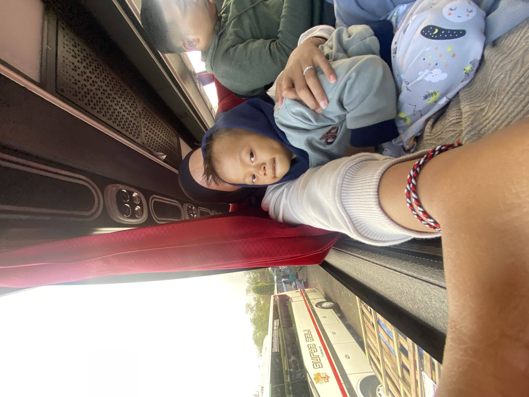 A young child is being held by an adult on a bus, with a red curtain to the left and a man in a green hoodie sitting behind them. The child is wearing a blue hoodie and a grey sweatshirt, and the adult is wearing a white sweater. Outside the window, another bus is parked, along with trees and other vehicles, suggesting an outdoor location, possibly a bus station or parking area in Melaya, Indonesia. The image is taken from the perspective of someone on the bus, looking out the window and at the people on board.