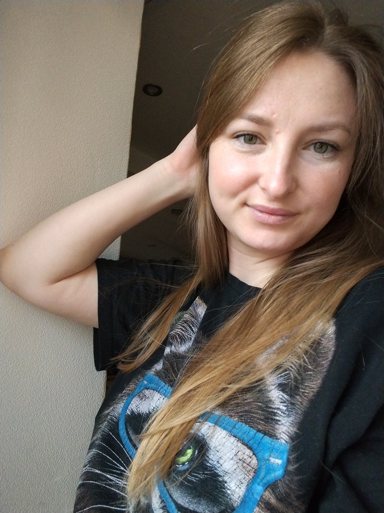 A young, light-skinned woman with long, light brown hair is leaning against a light-colored wall. She is wearing a black t-shirt with a graphic design featuring an animal (possibly a cat or tiger) wearing blue-rimmed glasses. Her right arm is raised, with her hand near her head. There is a doorway or opening visible in the background, leading to a darker space with a ceiling light. There is no information in the image to suggest that the image was taken in Chaikovsky, Russia.