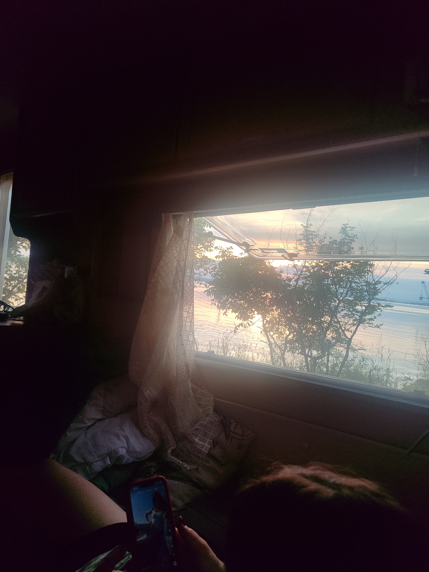 A dimly lit interior view from within a recreational vehicle or camper shows a person lying in bed, using a smartphone. The phone screen displays an image of a person. A sheer curtain hangs by an open window revealing an exterior view of water, possibly a lake or bay, illuminated by the setting sun. Trees and vegetation are visible outside the window. Based on the provided prompt, it is presumed the location is near Burgas, Bulgaria, and the body of water is part of the Black Sea.