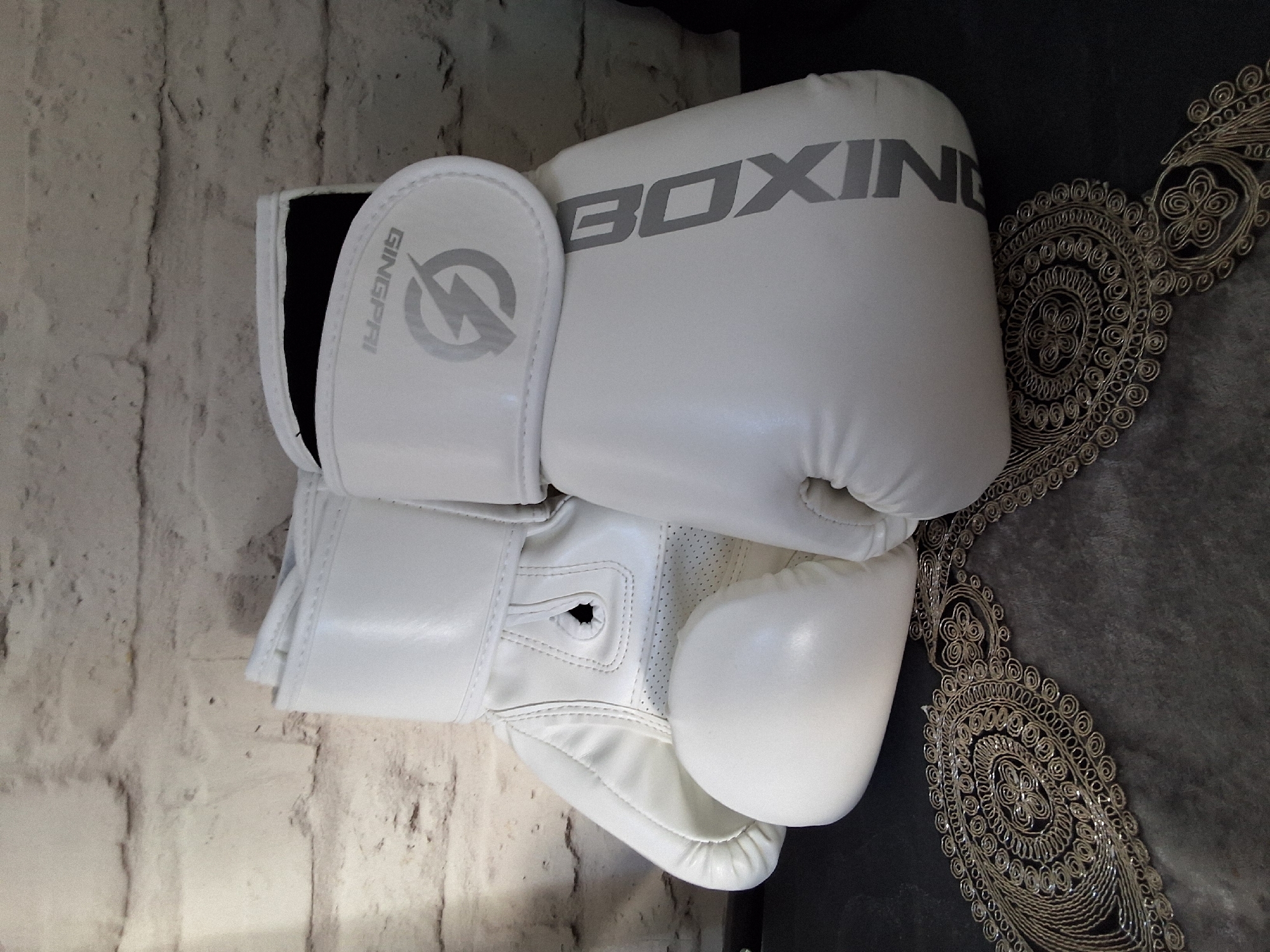 Two white boxing gloves are displayed against a white brick wall. The glove on the left is positioned slightly forward and to the left, with its palm facing upwards and its thumb curled inward. The glove on the right is placed behind and to the right of the first glove, also with its palm facing upwards and thumb curled. A black fabric lining is visible inside the cuff of the right glove. Both gloves appear to be new and are made of a shiny material. The right glove has gray lettering that reads "BOXING" vertically down its side, and a gray lightning bolt logo with the brand name "GINGPRI" above it. The gloves rest on a dark gray textured surface, possibly fabric, which is decorated with an ornate, swirling pattern of gold thread. The background is a light-colored, textured brick wall. No people or actions are depicted.