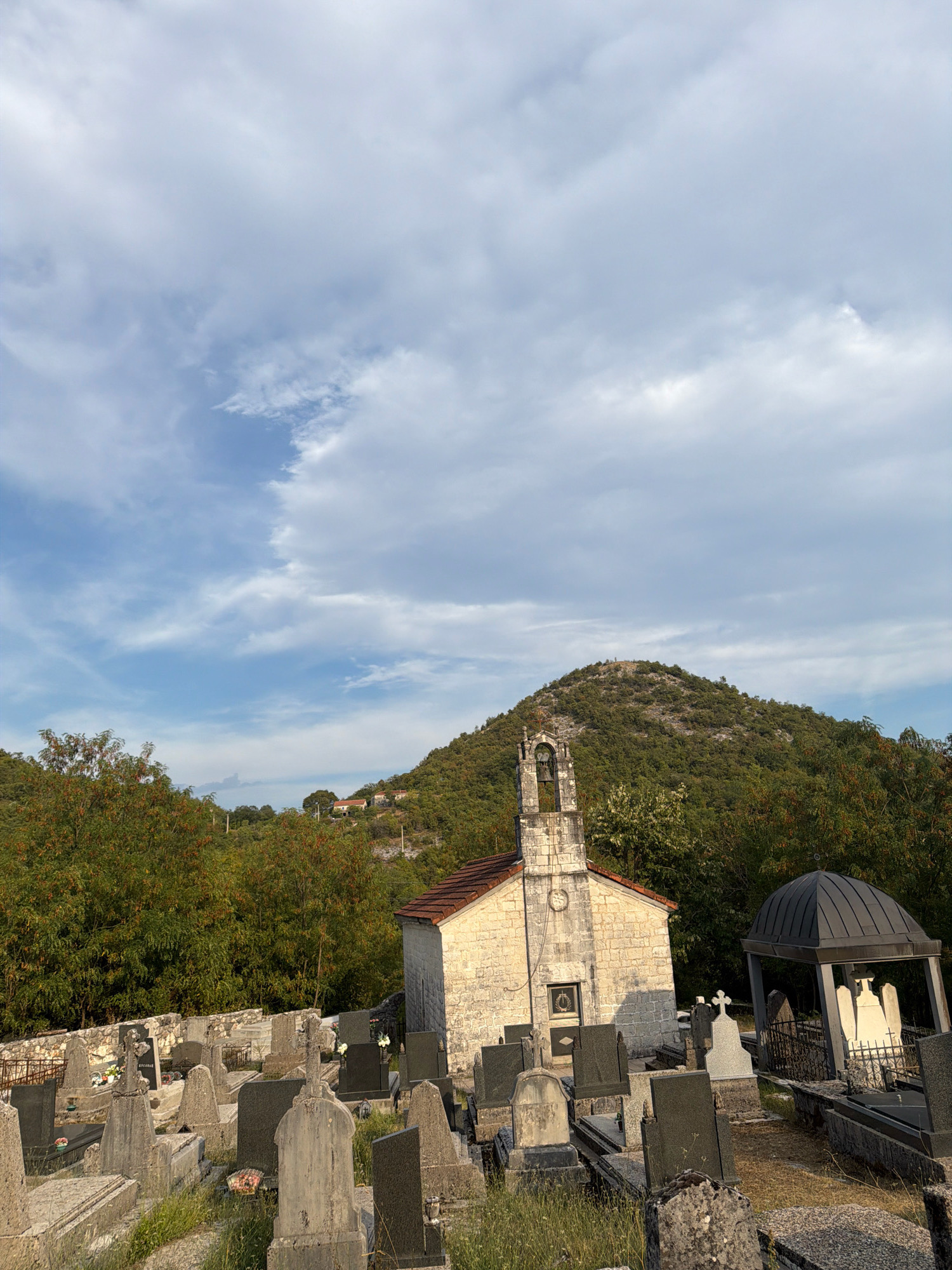 The image depicts a cemetery in Začir, Montenegro. In the foreground, numerous gravestones of various sizes and designs are visible, some with inscriptions and floral decorations. A small stone church with a bell tower stands to the right of the center. Adjacent to the church is a dark-domed mausoleum containing statues. The cemetery is surrounded by lush green trees and foliage. In the background, a verdant, tree-covered hill rises under a sky with scattered clouds. The scene appears to be set during daylight hours. There are no people visible in the image.