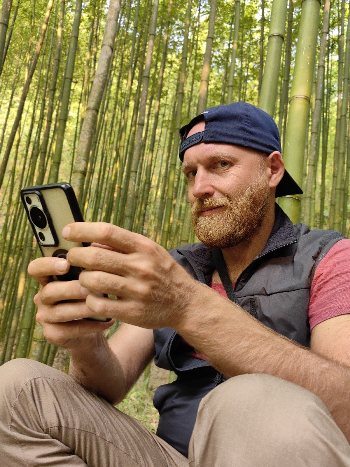 A male individual is depicted sitting in a dense bamboo forest in Mù Cang Chải, Vietnam. He is observed from the waist up, wearing a dark blue baseball cap backward, a dark gray zippered vest over a red t-shirt, and light brown or khaki pants. The man has a reddish-blond beard and mustache, blue eyes, and is looking towards the viewer with a slight smile. He is holding a light-colored smartphone with a dark protective case and a black circular grip on the back, with both hands positioned as if actively using the device. The background consists entirely of numerous tall, slender green bamboo stalks reaching upwards, indicating a natural, forested environment.