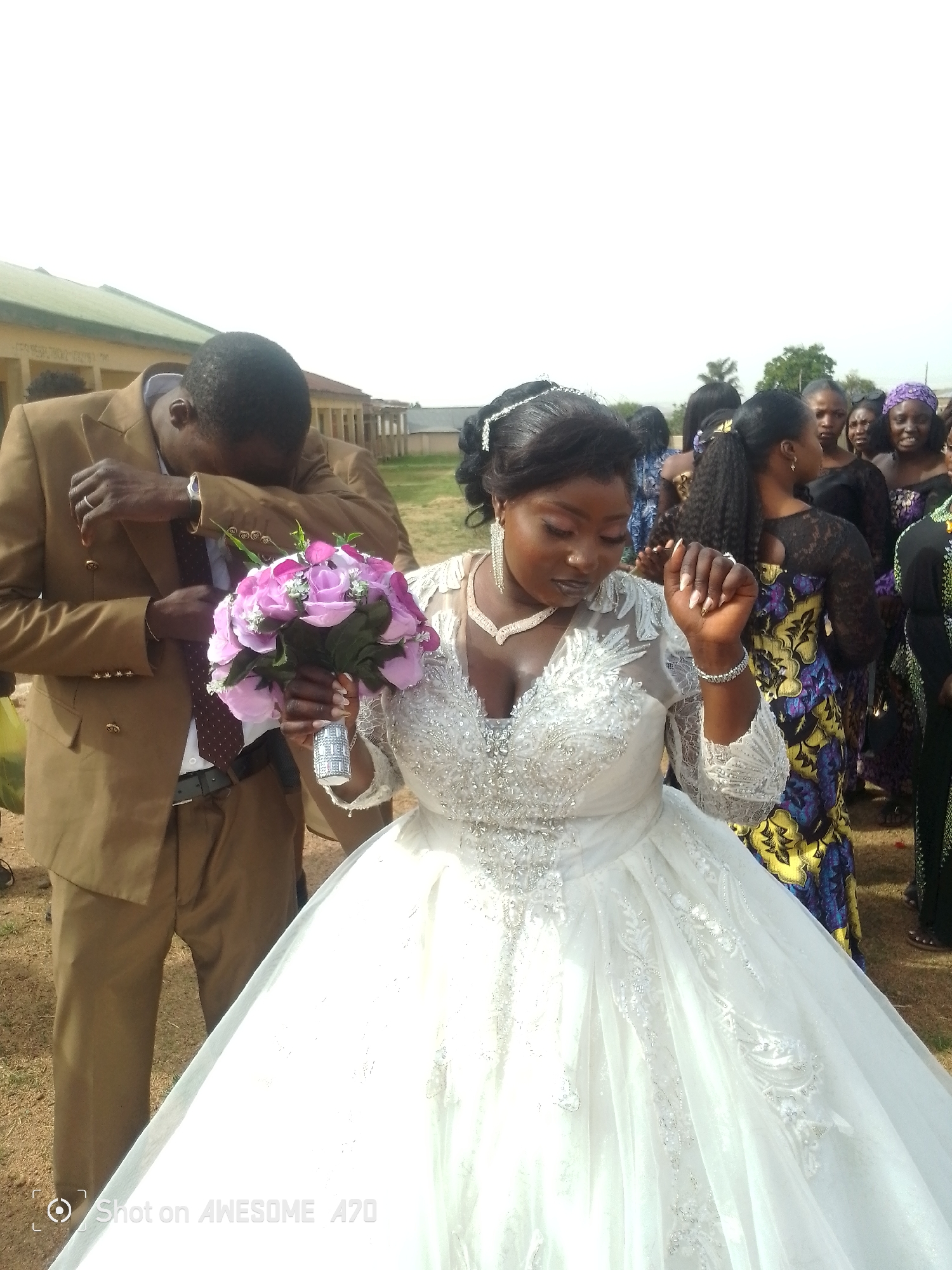 A wedding scene in an unidentified city. The bride wears a white gown with a tiara, necklace, and bracelet, holding a bouquet of pink and white flowers. A man in a brown suit stands to her left, covering his face with his arm. Several women dressed in colorful attire are visible in the background. The setting appears to be outdoors on a grassy area. The picture has the text "Shot on AWESOME A70" watermarked on the bottom.