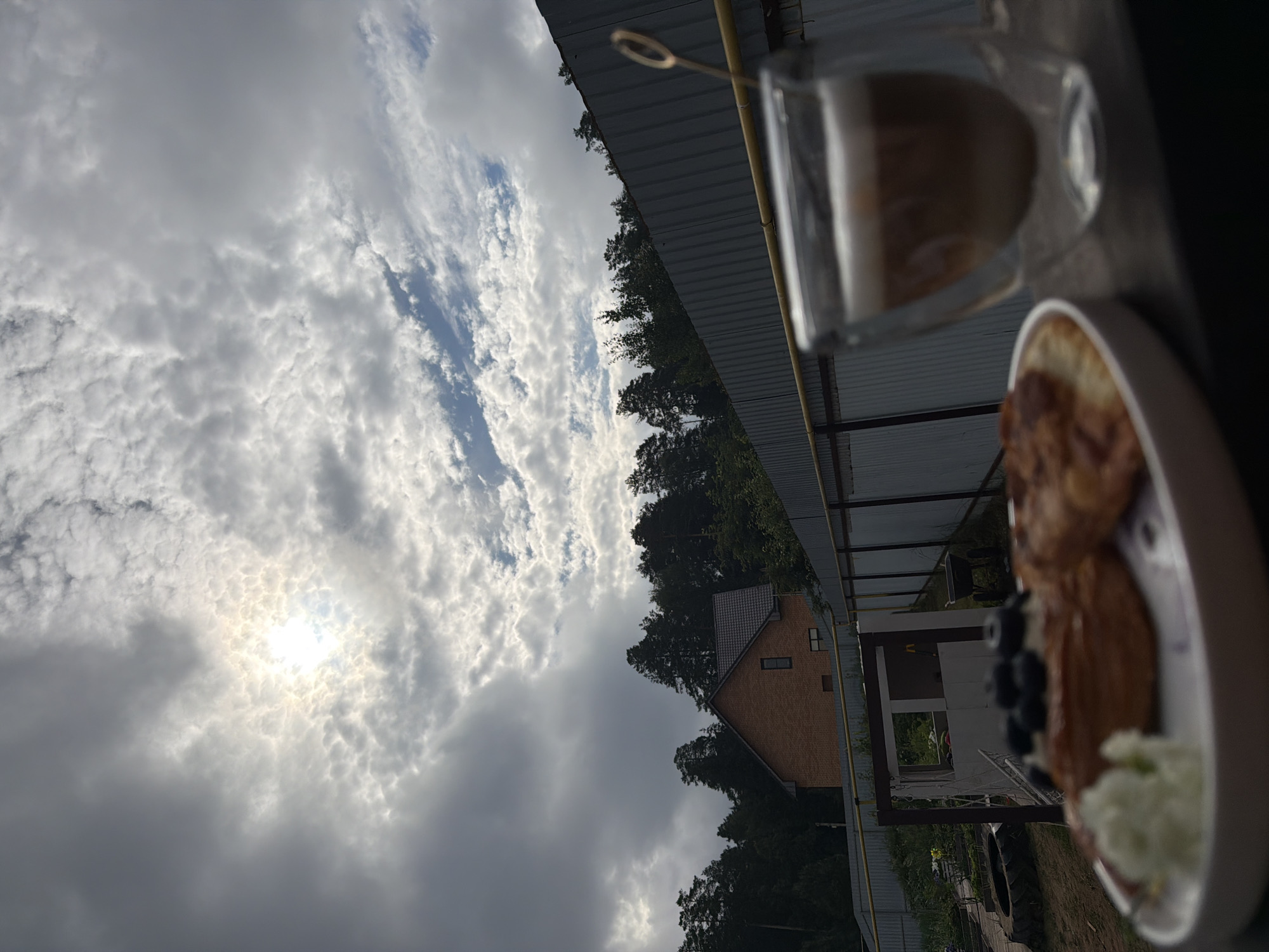 The image shows an outdoor scene with a breakfast spread in the foreground and a house and trees in the background under a cloudy sky. In the foreground, a white plate holds pastries topped with blueberries and a dollop of white cream. To the right of the plate is a clear double-walled glass mug filled with a layered coffee drink, likely a latte or cappuccino, with froth on top. A golden stirrer is visible in the mug.
The background features a corrugated metal fence running horizontally across the frame. Beyond the fence, a two-story brick house with a dark tiled roof is visible, surrounded by dense green trees. The sky is predominantly overcast with dramatic clouds, but the sun is breaking through in the upper left portion of the image, creating bright patches and illuminating the clouds. To the left of the house, there are indications of a yard with some structures and objects, including a tire and a small building. The overall setting appears to be a suburban or rural residential area. The image is taken from a low angle, looking up towards the sky and the background.