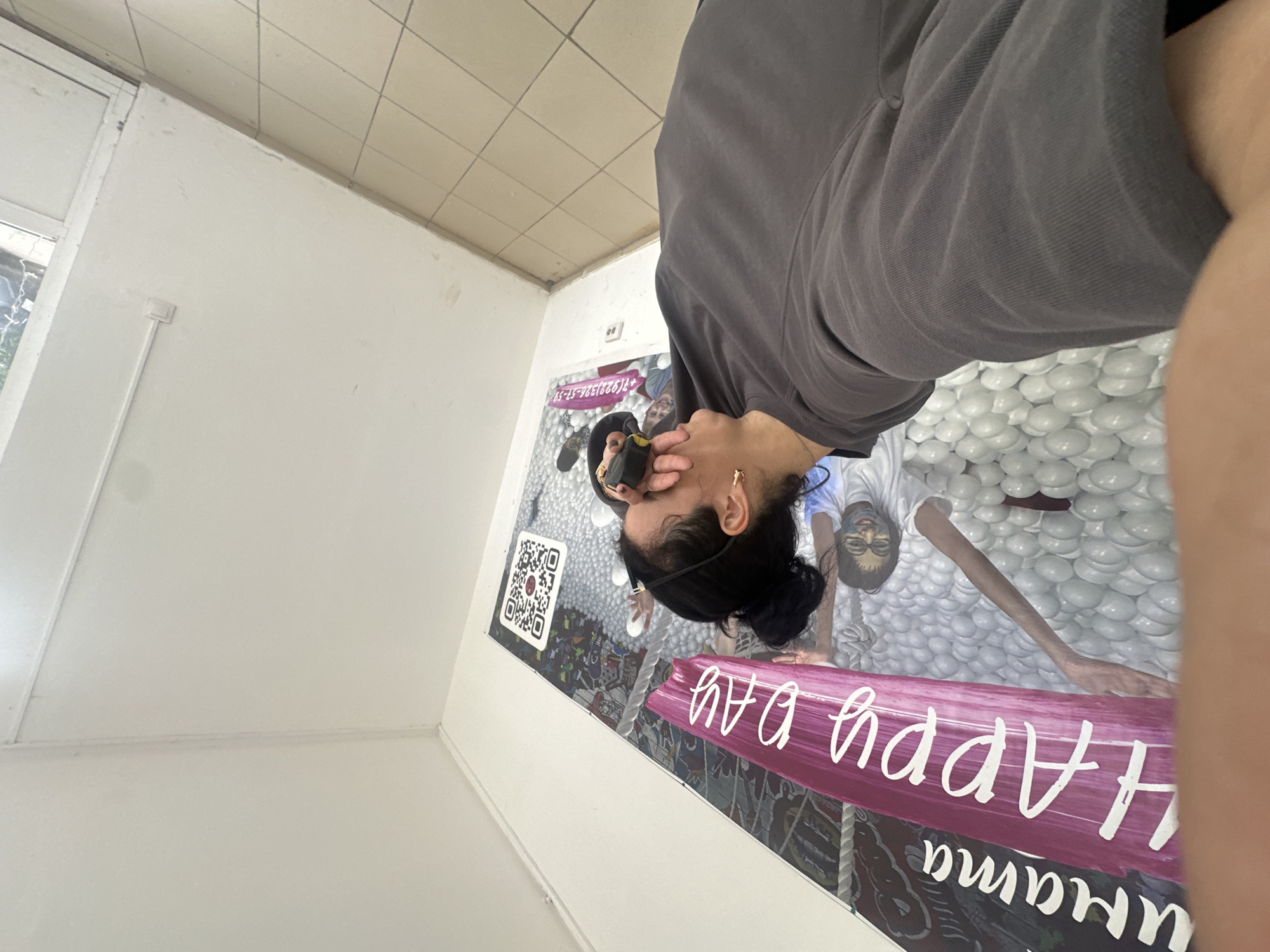 The image shows a person taking a selfie, or having their photo taken, in an indoor setting. The person is wearing a grey t-shirt and has dark hair pulled back. They are holding a yellow object up to their face, possibly a phone or a camera. In the background, there is a large mural or artwork on a white wall. The artwork features images of people and a lot of white, rounded shapes, resembling balls. There is also a QR code visible on the artwork. A large, pink banner with white cursive text is also part of the artwork. The text on the banner reads "Happy Mama". The overall scene appears to be a casual, possibly artistic or recreational space, like a gallery or an entertainment venue. The lighting suggests it is daytime, but the specific time or weather conditions are not discernible. The person's expression is difficult to read due to the angle and the object held to their face.