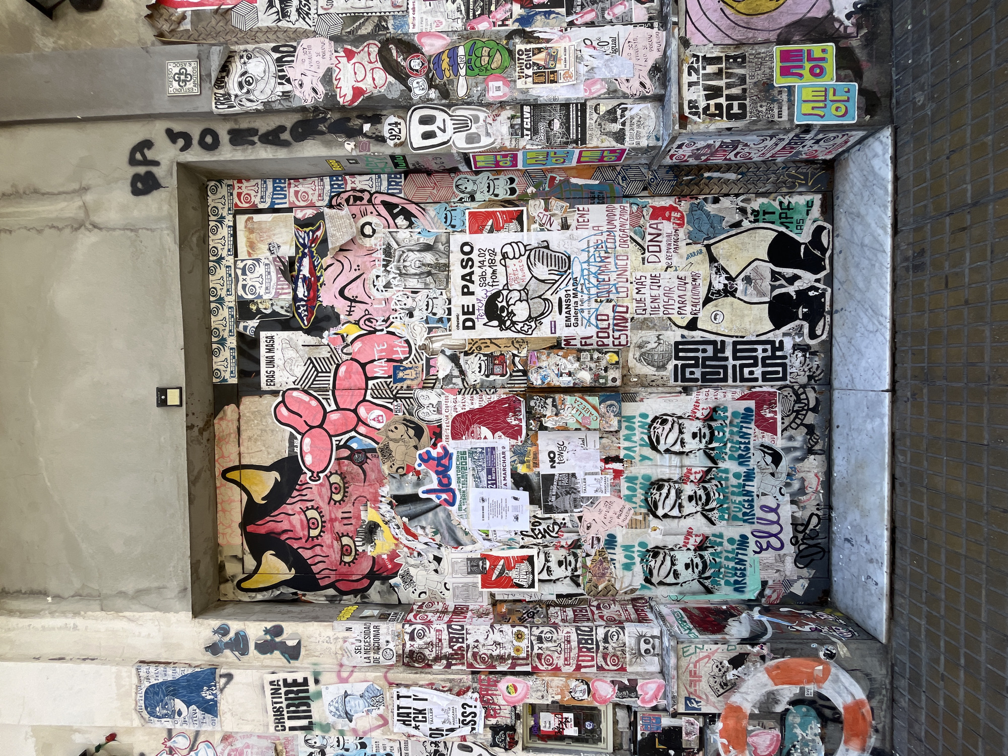This vivid image captures a heavily stickered and graffitied urban recess, likely a doorway or passageway in Buenos Aires, Argentina. The scene is dominated by a dense, layered collage of street art that covers the walls from floor to ceiling, creating a rich tapestry of colors, characters, and messages.
The subjects are primarily graphic art: countless stickers, stencils, and graffiti tags, featuring a wide array of stylized faces, creatures, and abstract designs. Notable characters include a large pink and red creature with multiple eyes and a skull-like motif, a small white skull with large eye sockets, a figure holding a mallet, and various cartoonish and distressed faces. The color palette is vibrant, with reds, pinks, blues, and black contrasting against the faded concrete and tiled surroundings.
The activity depicted is the cumulative result of urban expression and public art, suggesting a vibrant and active street art culture. No people are visible, emphasizing the art itself.
Notable details include the layered nature of the stickers, some peeling or torn, indicating a history of accumulation. The time of day appears to be daytime, with soft, even lighting. Location cues are abundant through the Spanish text. Visible text, both from OCR and visual inspection, includes: "CRISTINA LIBRE" (a political slogan), "PUEBLO ARGENTINO ARGENTINO ARGENTINO" (referencing Argentina), "DE PASO" (an event), "QUE MAS TIENE QUE PASAR PARA QUE REACCIONEMOS" (What else needs to happen for us to react?), "NO SOY VIOLENTO, NO SE PORQUE MUERDO" (I'm not violent, I don't know why I bite), "EMANS91 Galería MADA", "ESTUDIO GIESSO & AROTOS", and various other event details, brand names, and general urban slang like "ERAS UNA MASA" (You were great/a mass). The overall impression is one of rebellious creativity and a dynamic public space.