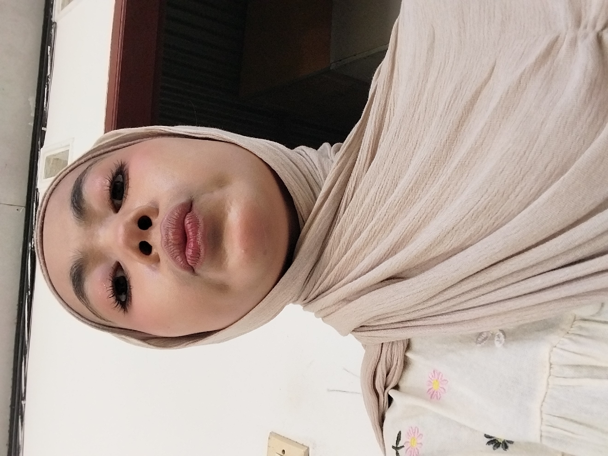 In Tibojong, Indonesia, a close-up, low-angle selfie shows a young woman wearing a light beige hijab and a cream-colored shirt with embroidered pink and black flowers. Her eyes are accentuated with pink eyeshadow and mascara, and her lips are pursed. The background is predominantly a white wall with a light switch on the left, and a dark, out-of-focus area on the right, possibly a doorway or furniture. The lighting appears to be from an overhead source, creating soft shadows. The overall impression is a casual, personal photograph taken indoors.