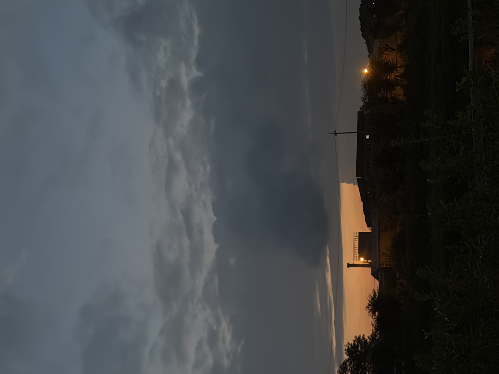 This is a vertical, low-angle, outdoor shot of a sunset or dusk scene in Dongducheon-si, South Korea. The sky is filled with dark, heavy clouds, with a sliver of orange light visible near the horizon. In the middle ground, there are several buildings with lights on, suggesting they are occupied. One building has a prominent chimney. The foreground is filled with dark silhouettes of trees and foliage. The overall impression is of a moody and slightly industrial twilight.