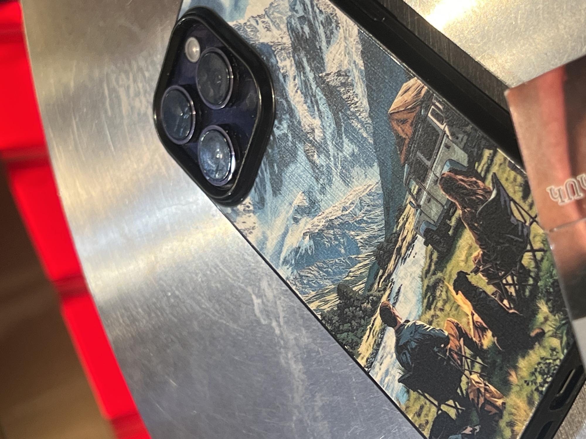 A high-angle, close-up shot displays a smartphone, possibly an iPhone, encased in a protective cover. The case is decorated with a detailed illustration depicting a mountainous landscape, complete with figures sitting in camping chairs and a vehicle. The camera housing of the phone is black and features three distinct lenses. The phone rests on a metallic surface. To the left of the phone, there's a red, blurred area. A portion of what appears to be a commercial package with text in Armenian is visible to the right of the phone. The scene is indoors. There is no information from the image that places it in Yerevan, Armenia.
