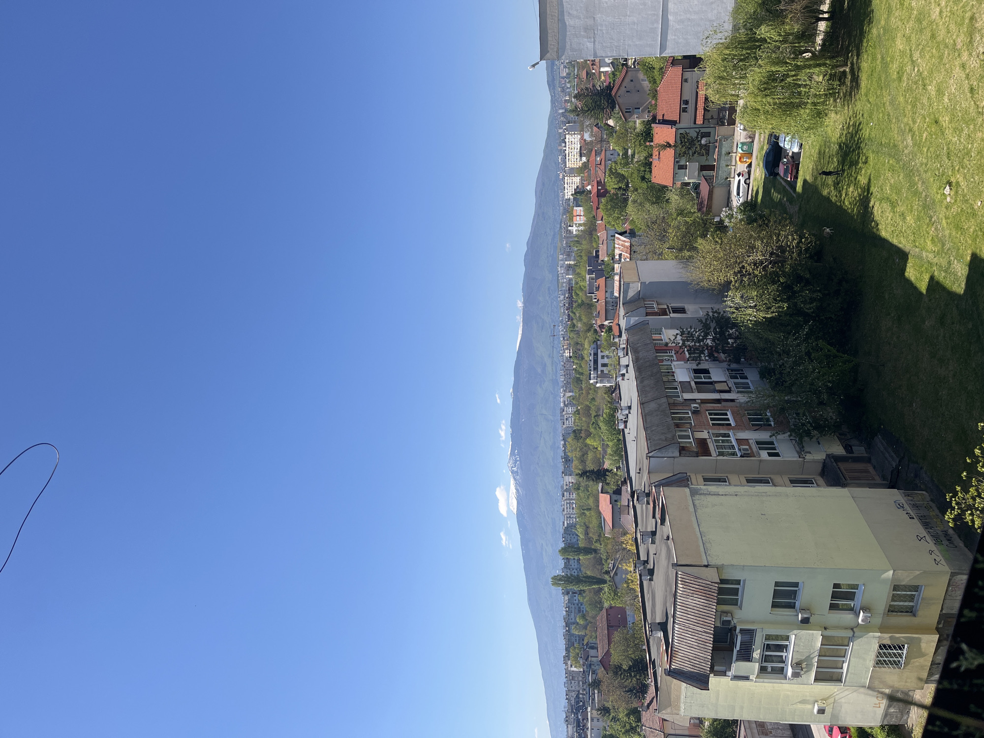 A high-angle, full daylight photograph captures a cityscape against a clear blue sky. The scene appears to be taken from a high vantage point, offering a view that includes buildings, a mountain range, and green spaces. Buildings are visible in the foreground and midground, varying in size and architectural style, with some having red-tiled roofs. A prominent mountain range stretches across the horizon, partially covered with what appears to be snow or cloud cover near its peak. Green spaces, possibly parks or gardens, are located on the right, suggesting urban landscaping. Based on the architectural style, landscape, and mountain range orientation, this photograph likely depicts a neighborhood within Sofia, Bulgaria, with the Vitosha mountain range visible in the background. No people are distinctly visible. There is some writing on the wall of a building, although the text is illegible due to distance and angle. Cars can also be seen parked near the green spaces.