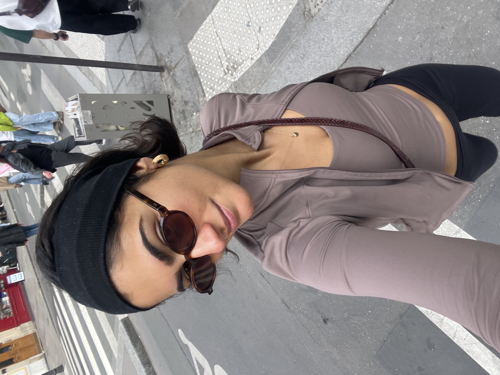 A medium-shot selfie shows a woman wearing a black beanie, brown round sunglasses, a gold earring, and a mocha-colored activewear jacket over a matching tank top. She is wearing black leggings and carrying a brown shoulder bag. The background shows a crosswalk with white stripes on a grey street in what appears to be a city. Other people are visible in the background, along with a metal object that appears to be a newspaper box. The scene is set in Paris, France, as identified by the presence of characteristic street furniture.