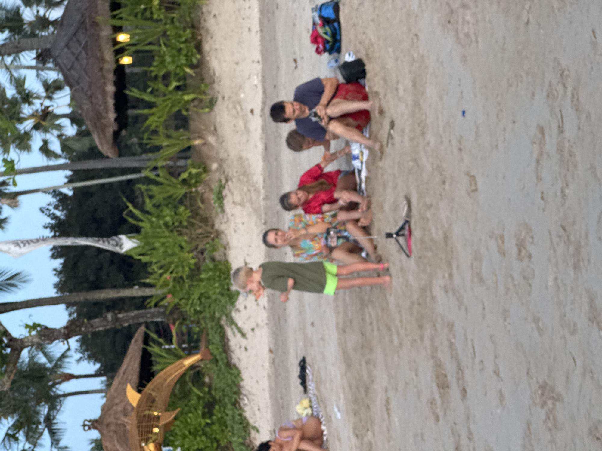 The image depicts a beach scene in Kuta Selatan, Indonesia, with several individuals and environmental elements.
**People:**
* A group of people are gathered on the beach, including adults and children.
* They appear to be sitting and interacting with each other.
**Environment:**
* The scene takes place on a sandy beach.
* Vegetation, including palm trees, is visible in the background, suggesting a tropical environment.
**Objects:**
* A towel or blanket is laid out on the sand.
* Beach gear or bags are present near the group.
**Actions and Interactions:**
* The group is sitting and interacting with each other, indicating social activity or relaxation.
* One person is standing and facing a group sitting down.