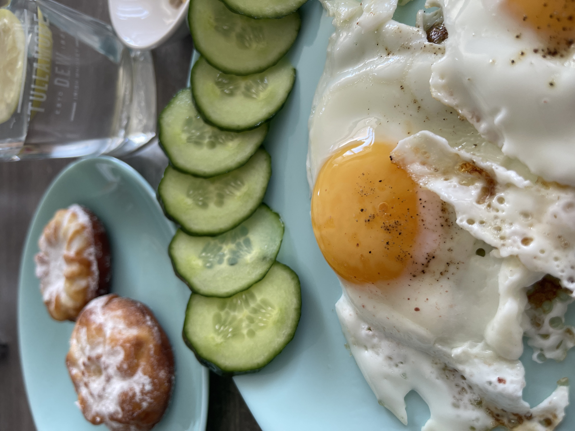 The image shows a breakfast meal on a table. In the foreground, there are two fried eggs with visible yolks and whites, seasoned with pepper. To the left of the eggs, there are several slices of cucumber arranged in a row on a light blue plate. In the background, on another light blue plate, are two small, round pastries dusted with powdered sugar. To the right, a glass filled with water and lemon slices is visible, with a dark surface in the background, possibly a table or countertop. A small white dish is also partially visible. The scene is brightly lit, suggesting daylight. The image does not contain any identifiable landmarks or features that would allow for its location to be determined as Blagoveshchensk, Russia.