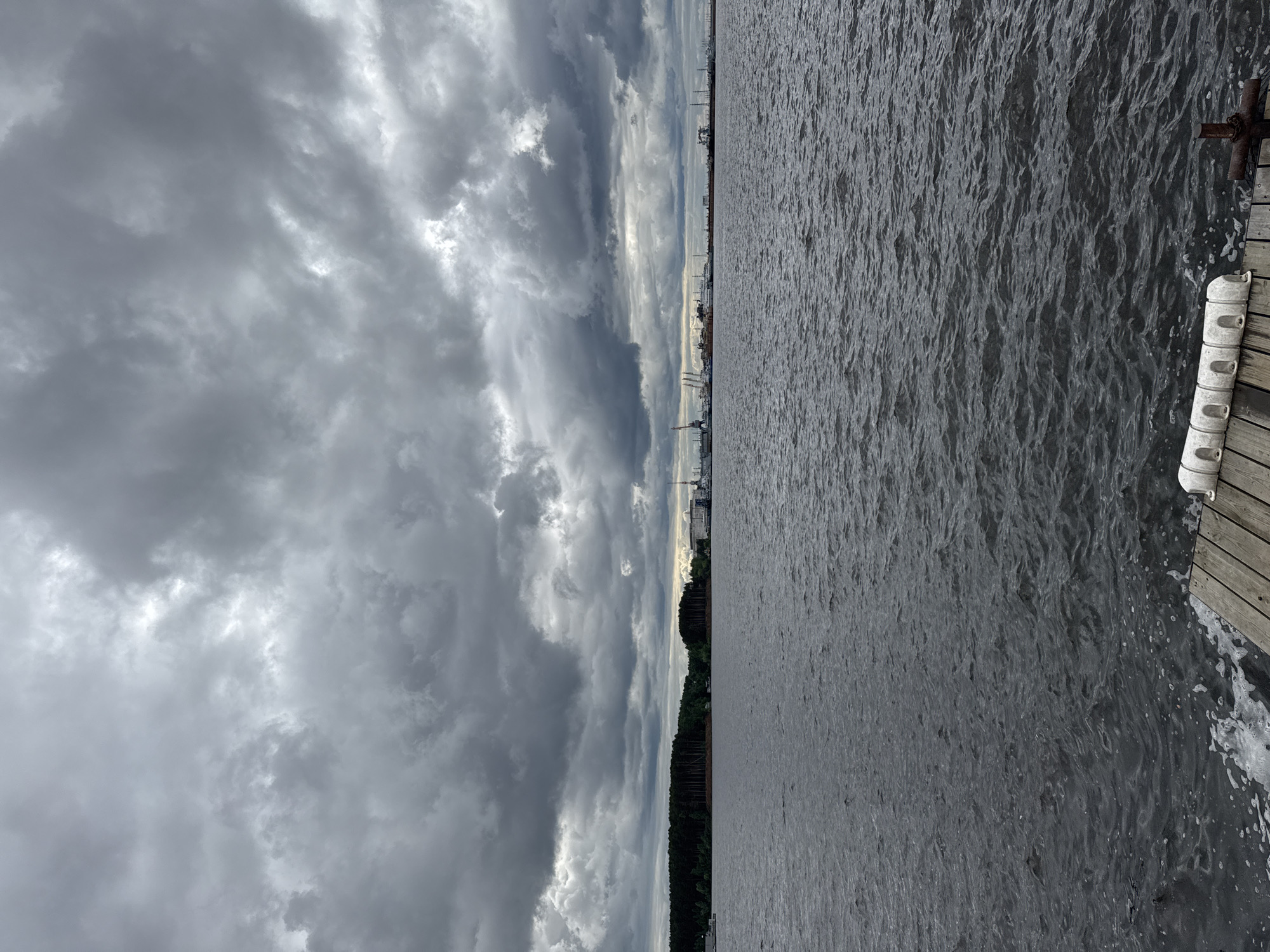 The image shows a waterfront scene in Chaikovsky, Russia. The perspective is from a dock, the edge of which is visible in the lower right corner with white cylindrical bumpers attached. The foreground features a body of water, likely the Kama River, with a slightly choppy surface. On the horizon across the water, the city of Chaikovsky is visible with industrial structures, cranes, and buildings silhouetted against the sky. The sky is filled with dense, gray storm clouds, indicating overcast weather. A line of trees is also visible along the shore.