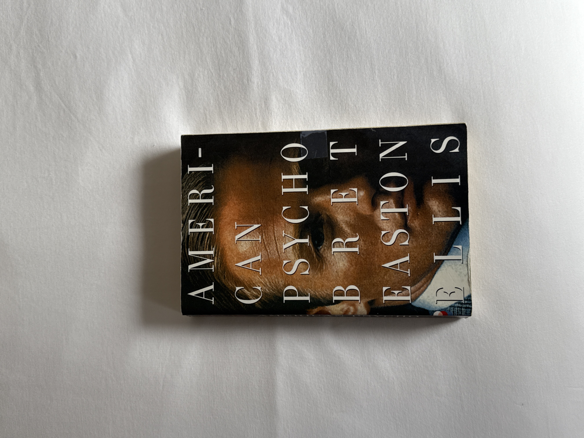 A paperback copy of the novel "American Psycho" by Bret Easton Ellis is depicted lying flat on a white, subtly textured fabric surface. The book is oriented horizontally, rotated 90 degrees clockwise, so its spine is towards the bottom of the frame and the cover text reads vertically from bottom to top. The cover features a dark background with a close-up of a man's face, presenting a neutral or intense expression. The man has light skin and short, dark hair, and is wearing a collared garment.
The title "AMERICAN PSYCHO" is prominently displayed in large, white, sans-serif capital letters across the top section of the cover. Below the title, the author's name, "BRET EASTON ELLIS," is printed in smaller, white, sans-serif capital letters. A small dark label or piece of tape is visible on the upper part of the cover, partially obscuring the word "PSYCHO." The white fabric surface exhibits faint wrinkles and creases, and the scene is lit evenly, creating a soft shadow directly beneath the book. The book itself shows slight wear along its edges.