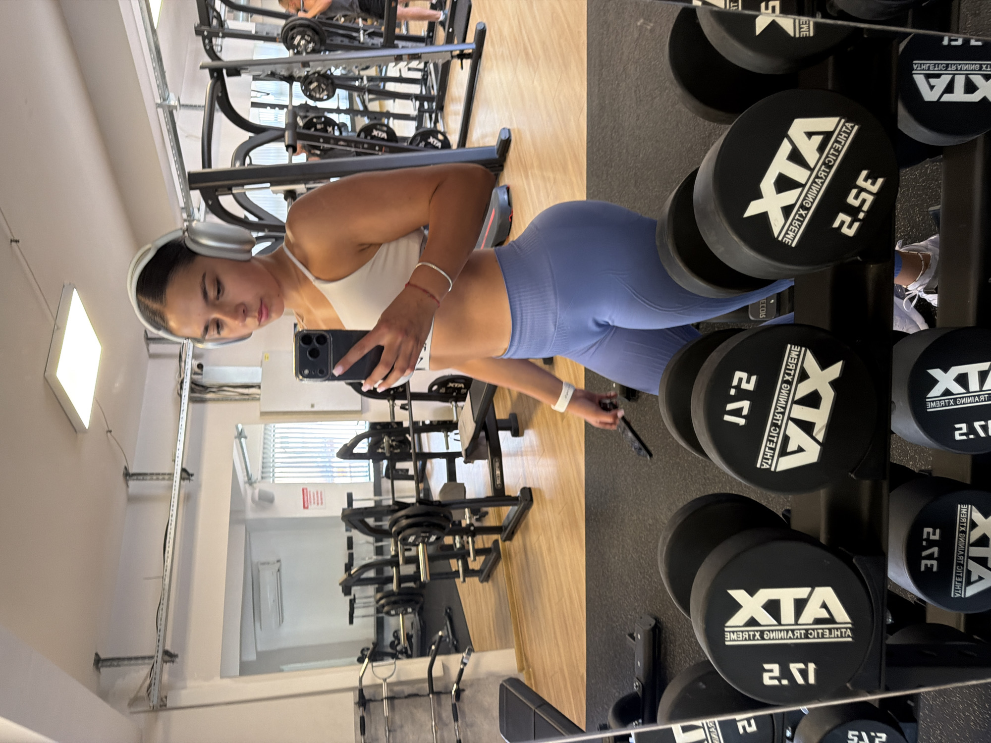 The image depicts a young woman in athletic wear taking a mirror selfie in a gym. She is wearing a white sports bra and light blue leggings. She has on silver headphones and is holding a smartphone in her right hand. The gym environment is visible in the background, with weightlifting equipment, including dumbbells and a bench press machine. The flooring appears to be a combination of wood and black matting. The lighting suggests an indoor setting, possibly during the day. The scene is clean and organized, indicating a functional workout space. There is no indication of the city of Gabrovo, Bulgaria, in the image.