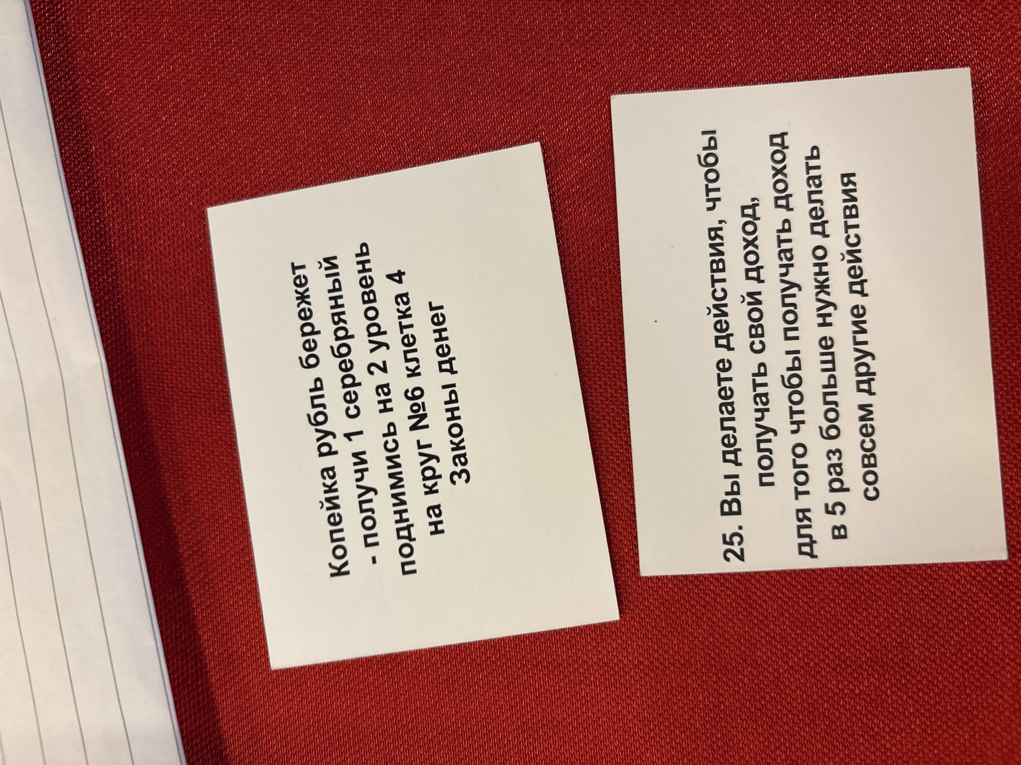 The image shows two small, white cards with printed text in Russian, placed on a textured, dark red surface. The card on the left reads: "Копейка рубль бережет - получи 1 серебряный поднимись на 2 уровень на круг №6 клетка 4 Законы денег" (A penny saved is a penny earned - get 1 silver coin, go up 2 levels to circle No. 6, cell 4. Laws of money). The card on the right reads: "25. Вы делаете действия, чтобы получать свой доход, для того чтобы получать доход в 5 раз больше нужно делать совсем другие действия" (25. You are taking actions to earn your income, to earn 5 times more income you need to take completely different actions). A notebook or notepad with lined paper is partially visible on the left edge of the frame. The image composition suggests a close-up shot on a tabletop or flat surface. There is no indication of the city of Kuta Selatan, Indonesia in this image.