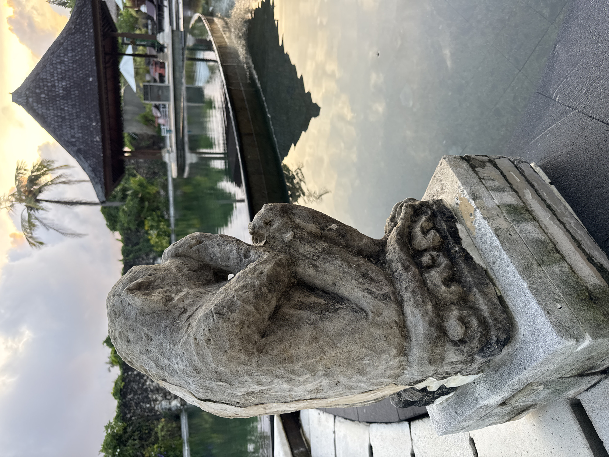 The image shows a stone statue, possibly of a mythical creature, placed on a stone pedestal near a pond or reflective pool. The scene appears to be in a tropical location, likely near a resort or spa. In the background, there's a building with a traditional Balinese-style roof. The reflections in the water suggest the presence of trees and sky, possibly at sunset or sunrise given the warm colors. Based on the location provided, the image is likely taken in Jimbaran, Indonesia, near a water feature within a resort or public space. The statue is worn and weathered, suggesting it's been exposed to the elements for some time. The pond has a curved bridge or walkway over it. The angle of the image is low, highlighting the statue in the foreground.