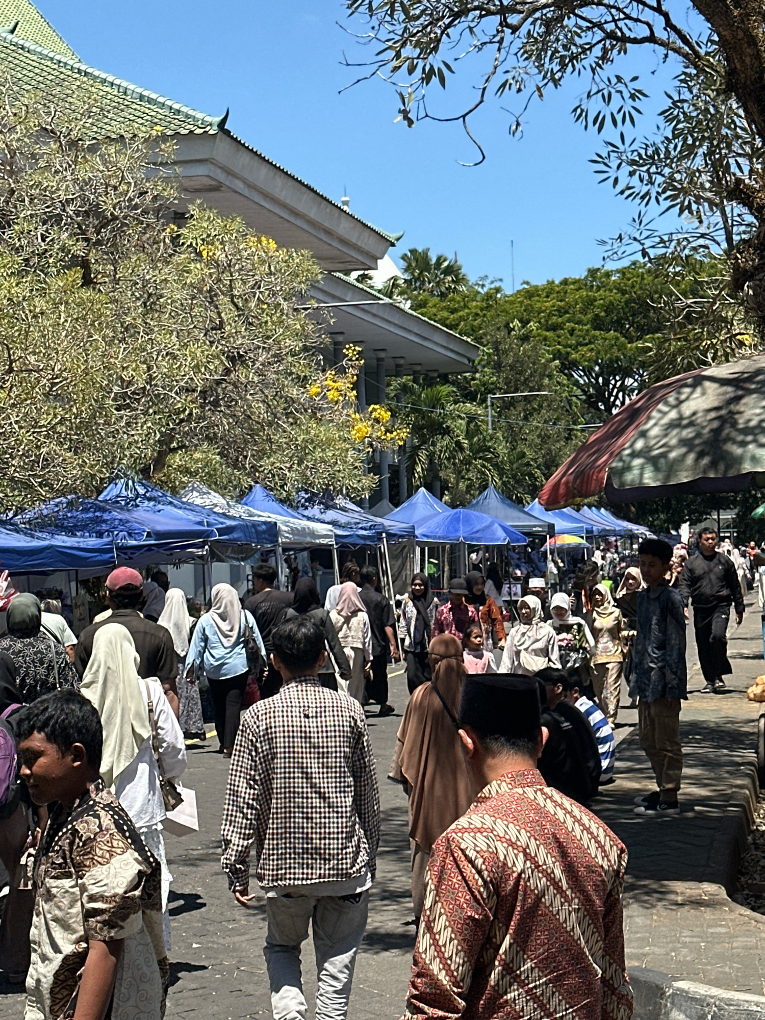 The image depicts a bustling street scene with numerous people walking past tents set up along the side of a road. The tents, predominantly blue and some with white canopies, appear to be stalls or market booths. The people are dressed in a variety of clothing, with many women wearing headscarves, indicating a cultural context. The architecture visible in the background, particularly the building with a green tiled roof and elaborate eaves, suggests a location in Malang, Indonesia, possibly near a significant cultural or governmental building. The atmosphere is bright and sunny, with a clear blue sky overhead and trees lining the street, some with yellow flowers. The ground is paved, and there is a slight elevation or curb along the side of the road where the tents are situated. The overall impression is of a lively outdoor market or event taking place in a city.