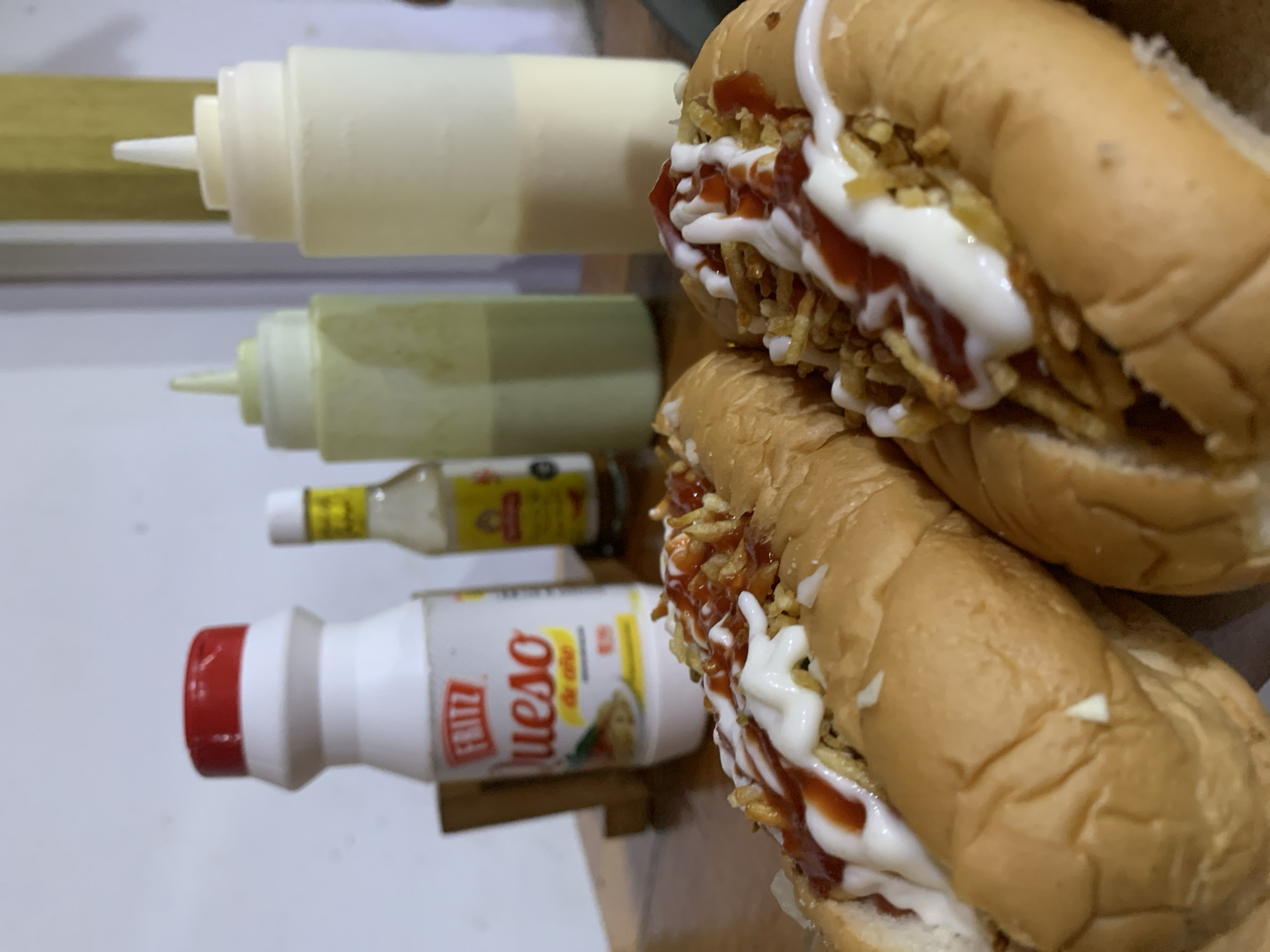 The image displays two generously topped hot dogs alongside a selection of condiment bottles on a light-colored surface, likely a table, in what appears to be an indoor setting in Santa Ana, Venezuela. The scene captures a prepared meal or snack with accompanying sauces.
The two hot dogs, presented in soft buns, are stacked on top of each other. Both are abundantly garnished with a layer of crispy, thin fried potato sticks (papas hilo), drizzled with bright red ketchup and white mayonnaise, creating an appealing contrast of colors and textures.
To the left of the hot dogs, four different condiment containers are visible. From top to bottom:
1. A large, opaque white squeeze bottle, likely containing mayonnaise or a similar white sauce.
2. A smaller, translucent pale green or yellowish squeeze bottle, possibly holding a guacamole-based sauce or another creamy condiment.
3. A small, clear glass bottle with a yellow label, suggesting it contains a liquid sauce such as hot sauce or vinegar.
4. A white plastic bottle with a prominent red cap and a red "FRITZ" logo on its side. Below the logo, the words "Queso de" are visible, indicating it contains cheese sauce, likely for the hot dogs.
The overall impression is a casual and popular fast-food style meal, characteristic of "perros calientes" often found in Venezuela. No people are visible, and the time of day is not discernible, but the lighting suggests an indoor environment.