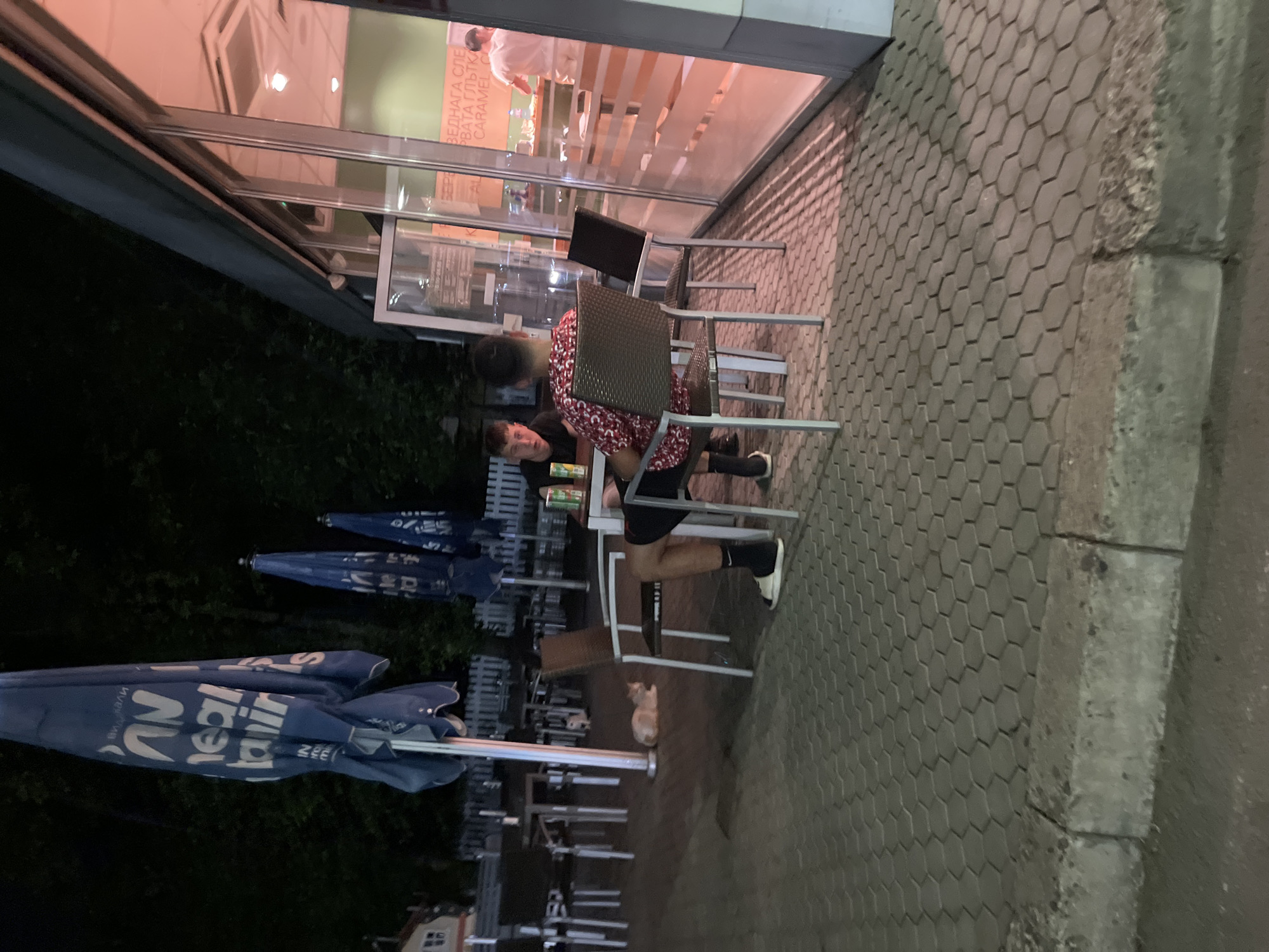 A man and a woman are seated at a table outside a building with glass windows. The woman wears a red and white patterned shirt, while the man is in black. An open tin can is on the table. The surrounding area includes outdoor seating with blue umbrellas, and a light-colored cat is nearby. The location is in Popovitsa, Bulgaria.
