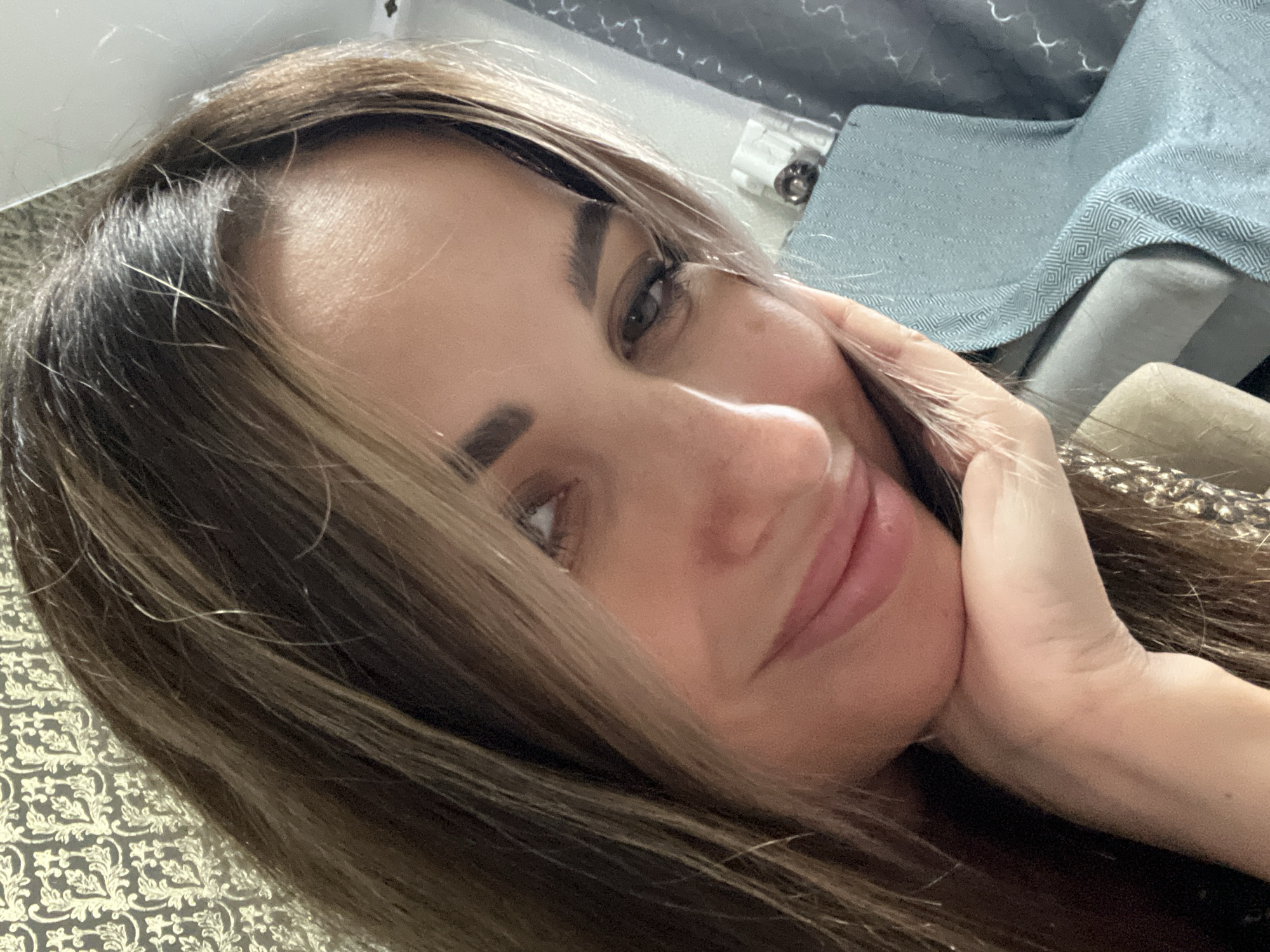 A close-up, selfie-style photograph shows a woman's face in profile, with her right hand supporting her chin. She has long, layered brown hair with blonde highlights. Her eyes are dark and looking slightly to the left, and her lips are slightly parted in a subtle smile. She appears to be indoors, with a patterned wallpaper visible in the background to the left and a blue textured fabric to the right, possibly a blanket or upholstery. There is also a small object, possibly a cup or container, visible on a surface behind her. The lighting suggests an indoor setting.