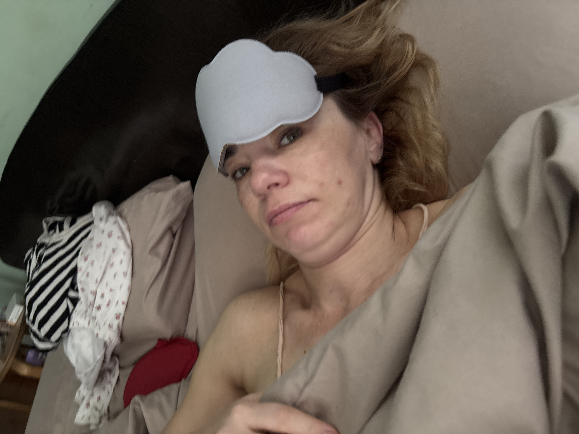 A woman lies in bed wearing a light blue sleep mask. She is covered by a tan comforter, and her right arm is visible, resting on the comforter. Behind her, a dark brown headboard is visible, and to her left, a pile of clothes, including a black and white striped shirt and a floral print shirt, are on the bed. The environment appears to be a bedroom, and the overall scene suggests the woman is preparing to sleep or has just woken up. The location is specified as Vsevolozhsk, Russia.