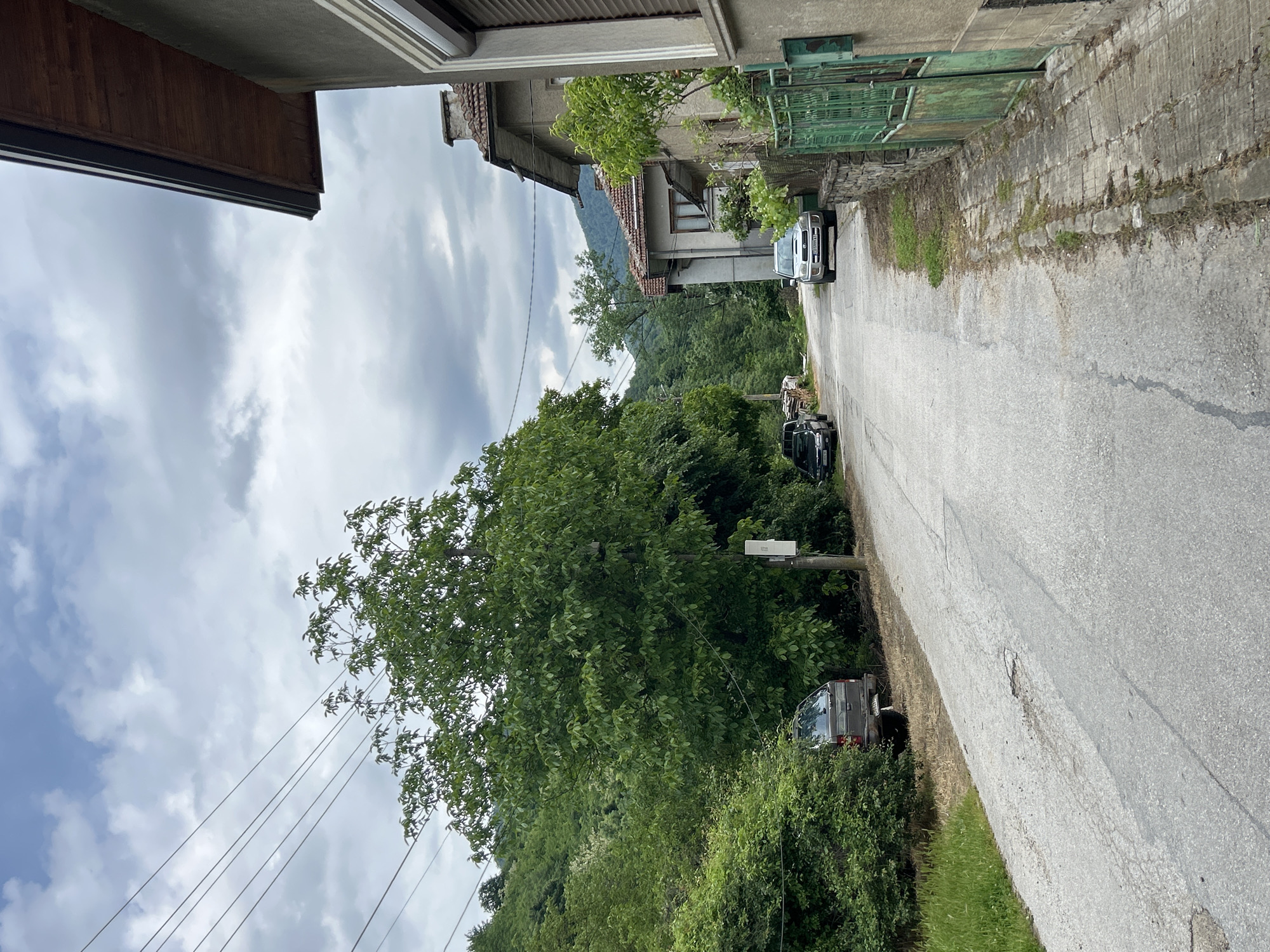 A street scene in an unknown location. The street runs downhill from right to left in the image. Three cars are parked on the sides of the street. Lush green vegetation covers the lower portion of the image. Utility poles with wires run along the left side of the street. Buildings are visible on both sides of the street, with a mountain visible in the background between buildings. The sky is overcast with heavy clouds.