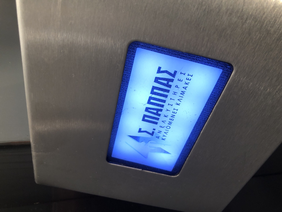 The image captures a close-up of an illuminated sign embedded within a brushed metal surface, likely stainless steel. The scene is set in Moschato, Greece. The rectangular sign has rounded corners and a distinct blue, textured border. It glows with a bright blue-white light, making the text and a logo prominent.
No people are visible, and no specific activity is taking place; the image focuses on the static installation. The metal surface shows subtle signs of wear, such as fine scratches and reflections.
The text on the sign is in Greek. The main name, prominently displayed, is "Σ. ΠΑΠΠΑΣ" (S. PAPPAS). Below this, in smaller capital letters, are two lines specifying services: "ΑΝΕΛΚΥΣΤΗΡΕΣ" (Lifts/Elevators) and "ΚΥΛΙΟΜΕΝΕΣ ΚΛΙΜΑΚΕΣ" (Escalators). This indicates that S. Pappas is a company dealing with lifts and escalators. Below the service description, a stylized logo is visible, appearing as an angular, abstract shape, possibly a stylized letter or a symbol like a lightning bolt.
The illumination of the sign suggests it is either an internal light source or captured in low light conditions, designed for clear readability. The object featuring the sign is likely a component of an elevator or escalator system, or an informational panel associated with such equipment.