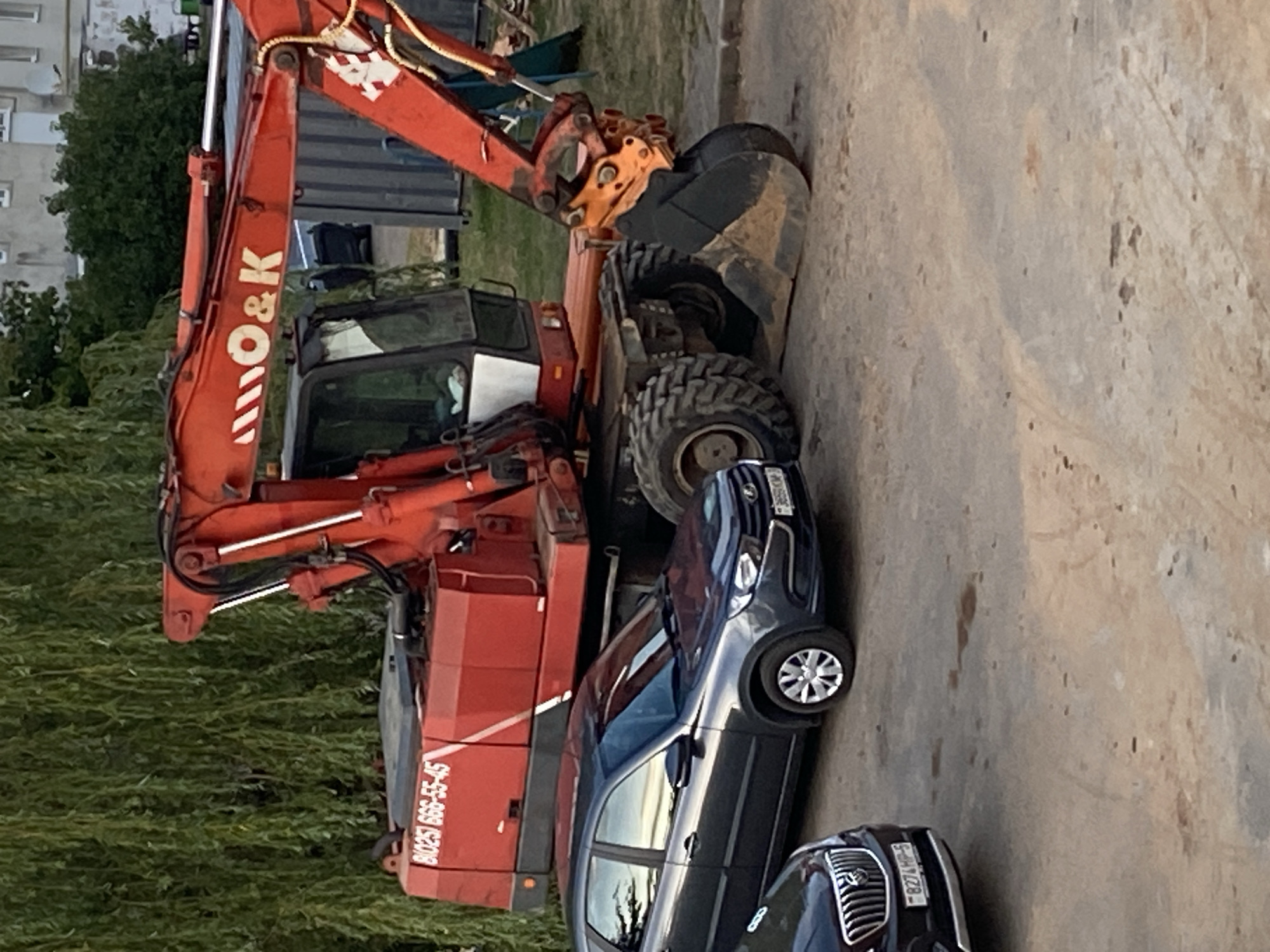An orange O&K excavator with the phone number B1(025) 666-55-45 is parked on a paved surface in Krupki, Belarus. A dark gray hatchback car is parked to the left of the excavator, and a portion of a dark-colored sedan is visible to the far left. The background includes a large weeping willow tree, a building, and a metal shipping container. The scene appears to be outdoors in a residential or industrial area.