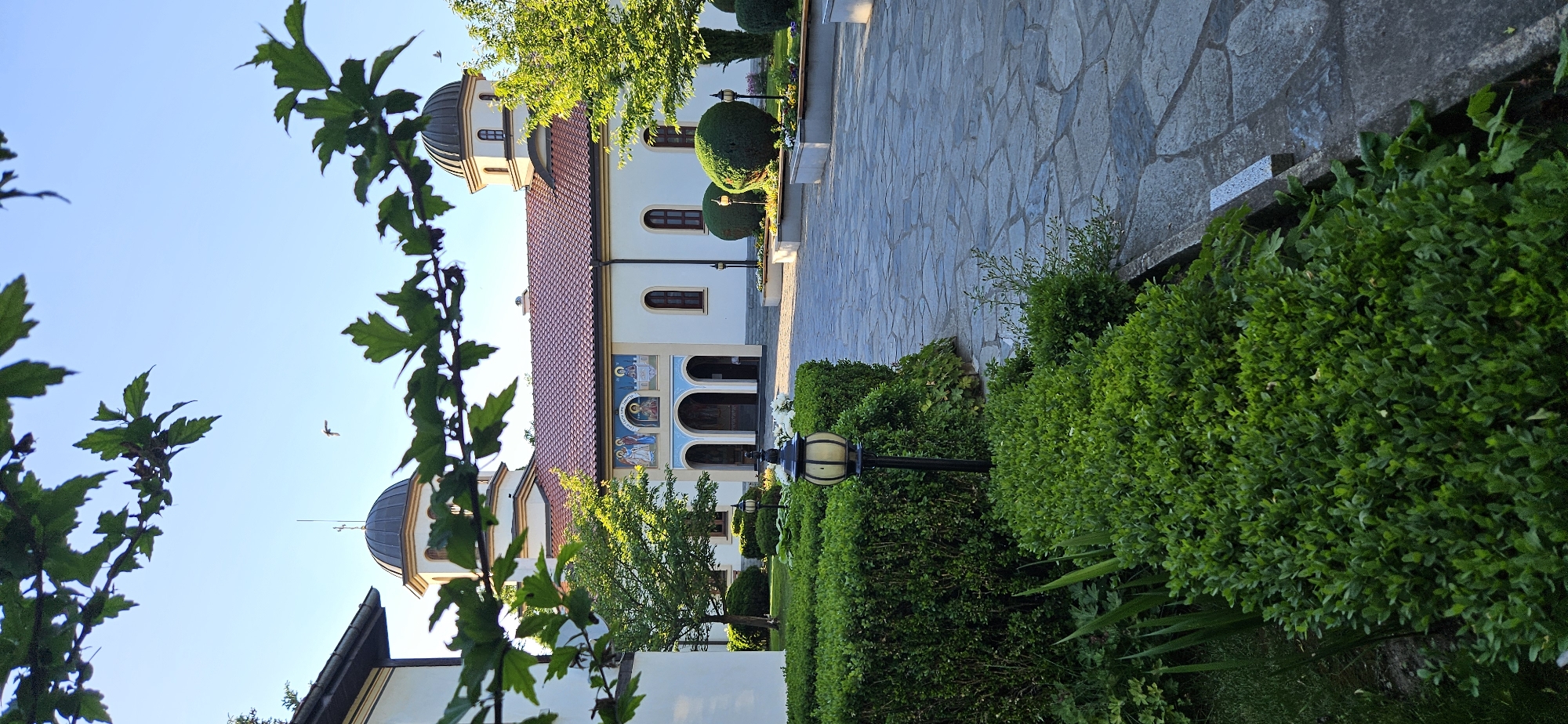 A street scene is depicted with a building, possibly a church, in Blagoevgrad, Bulgaria. The building has a red tiled roof, white walls, and arched windows. It features a dome structure and decorative artwork above the entrance. A stone walkway leads up to the building, flanked by manicured green shrubbery and trees. Street lamps are interspersed among the foliage. Branches and leaves frame the left side of the view, creating a natural foreground. The sky is a clear blue. A small bird is visible flying in the sky.