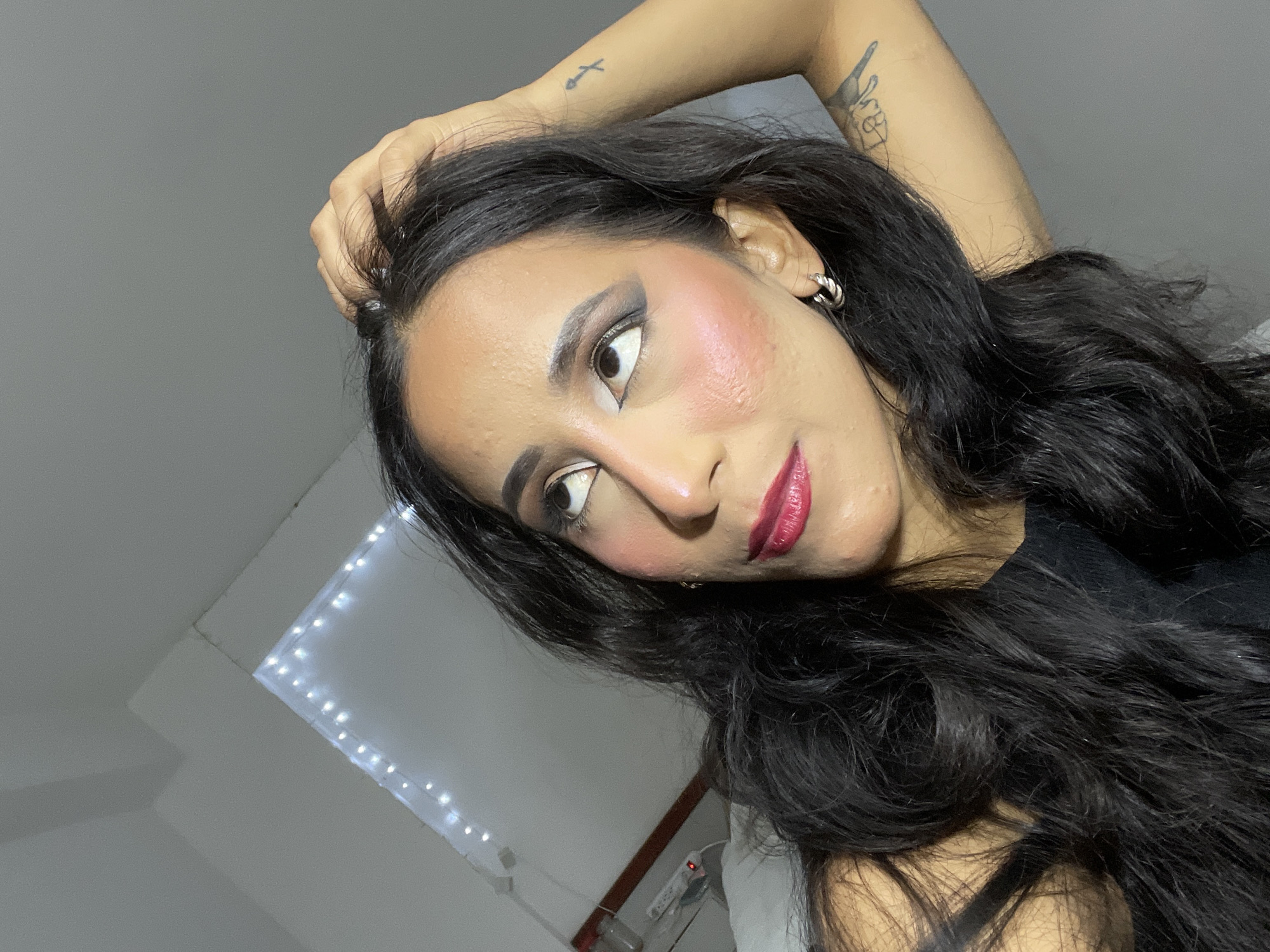 A medium shot captures a woman with dark, wavy hair, adorned with dramatic makeup, looking upwards with her right hand scratching her head. She is positioned in a room with white walls, a corner of which is decorated with string lights. The woman wears a black top and silver hoop earrings. Her left arm is bent, with tattoos visible on her right arm and upper left arm. The room appears to be indoors, possibly a bedroom, given the presence of a white bed with a pillow visible. There are also some electrical outlets and cables on the wall in the background. The overall lighting suggests it may be nighttime or a dimly lit interior space. The specific location within Porlamar, Venezuela, is not discernable from the visual information provided.