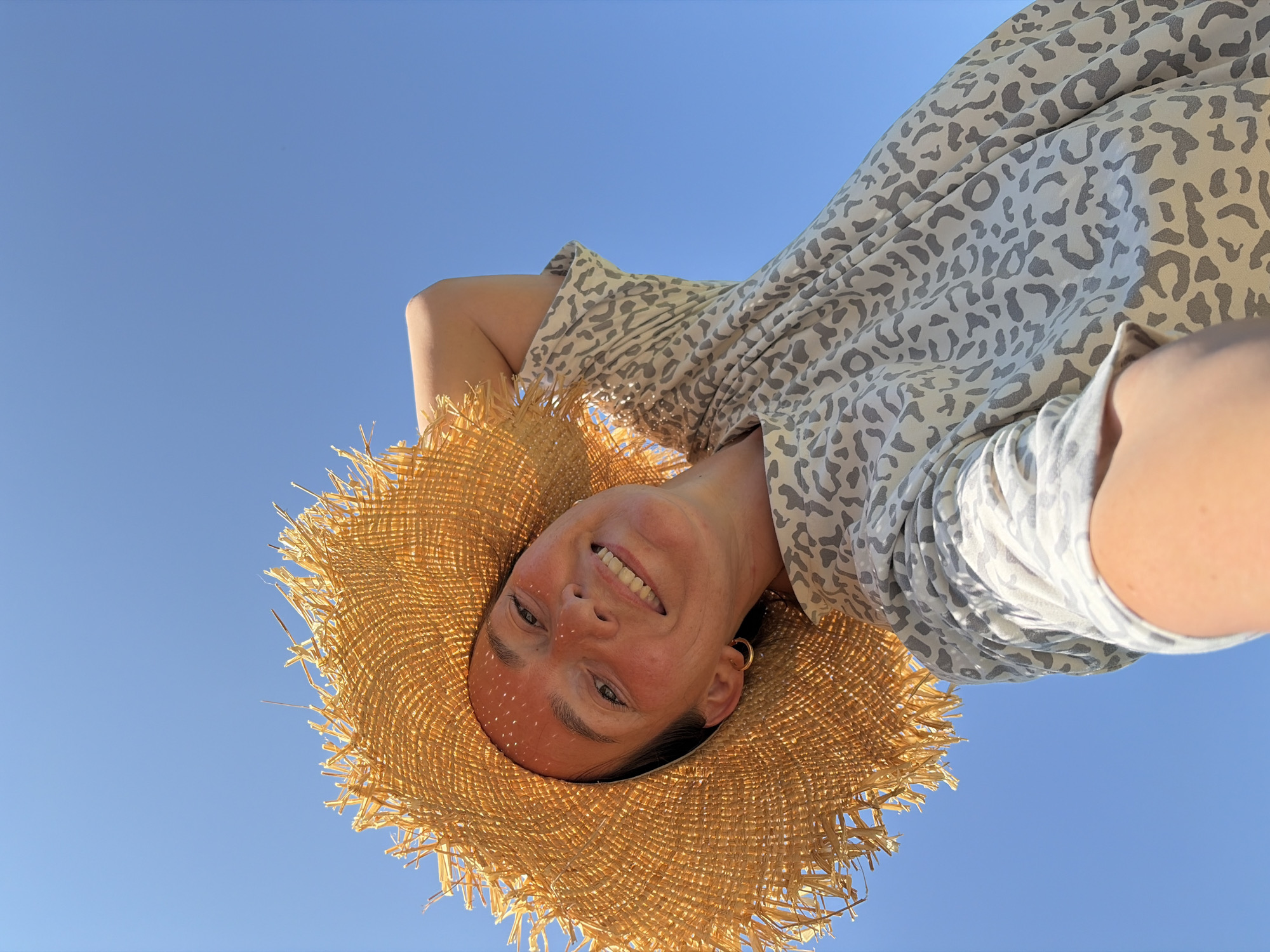 A woman is looking up at the camera, wearing a large straw hat and a patterned dress. She is smiling, with her left arm raised and resting on her head. The background is a clear blue sky. The image does not provide any information about the location in the city of Ольховка, Russia.