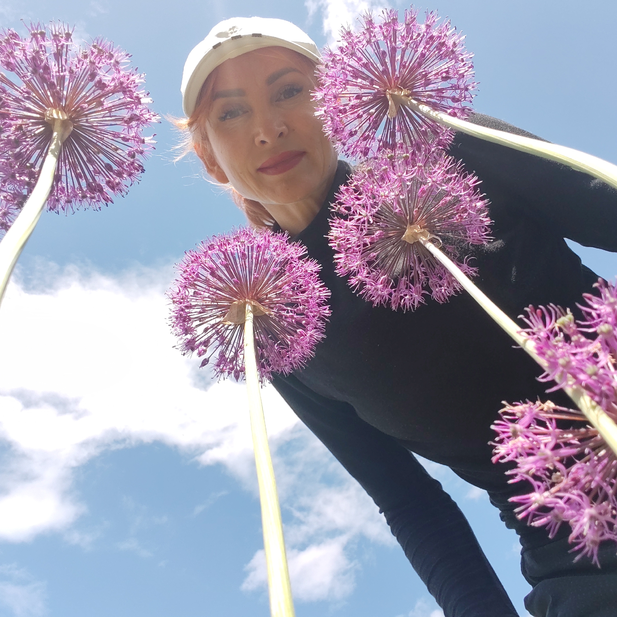 The low-angle image shows a person in a white cap and black long-sleeved shirt surrounded by several globe allium flowers. The person has fair skin, red hair, and red lipstick. The flowers are in full bloom and display a vibrant purple color. The sky is visible in the background with scattered clouds. The location is not discernible within the content of the image.