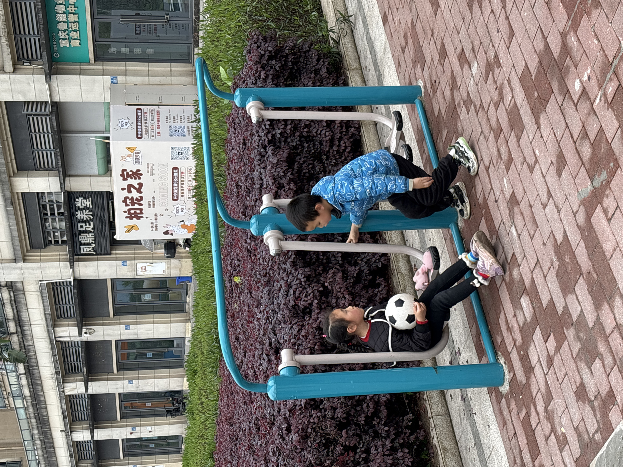 The image captures an outdoor urban scene in Jiangjin, Chongqing, China, featuring two children by public fitness equipment. In the foreground, a young girl wearing a black jacket and black pants sits on the foot pedals of a turquoise-and-white outdoor elliptical machine, holding a black and white soccer ball. A pair of pink slippers rests on the adjacent pedal. To her right, a young boy in a blue patterned jacket and black pants stands beside the machine, his right hand extended towards a component of the equipment. They are positioned on a reddish-brown brick pavement, which shows some wet patches. Directly behind the children and the equipment is a manicured hedge of dark red/purple foliage. In the background, a multi-story commercial building with a light-colored facade, dark windows, and horizontally slatted vents is visible. Storefronts include "凤鼎足养堂" (Fengding Foot Health Hall) and a large white banner for "拍宠之家" (Pai Chong Zhi Jia - Pet Home) with "COMING SOON" in English, partially obscuring a sign for "重庆鲁能物业 商业运营中心" (Chongqing Luneng Property Commercial Operations Center).