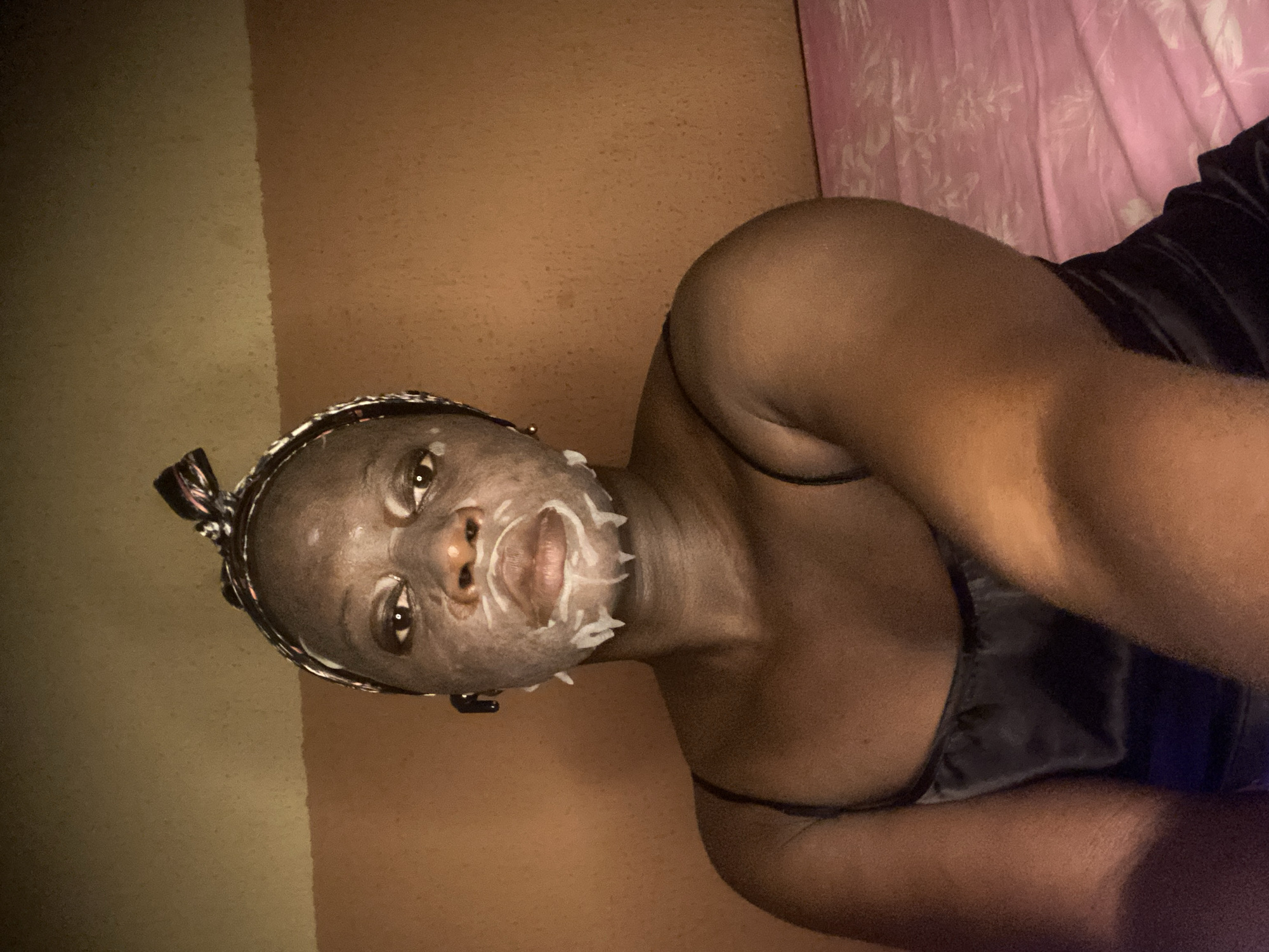 An indoor, medium shot captures a person of color, possibly female, lying down and looking towards the camera, suggesting a selfie or self-portrait. The subject has dark skin and is wearing a dark, possibly satin, camisole or top with thin straps. A greyish facial mask with white streaks, particularly prominent around the mouth and nose, is applied to their face. A decorative headband or braided hair accessory is visible around their head.
The person appears to be engaged in a self-care routine, possibly relaxing in a bedroom in Maigizo, Nigeria. The lighting is dim, suggesting it is evening or nighttime. The background on the left features a wall or surface with a two-tone color scheme, transitioning from a lighter brown to a darker, reddish-brown. On the right, a pink curtain or fabric with a floral or patterned design is visible, along with a dark blanket or sheet. The person's expression is calm and neutral. There is no visible text in the image.